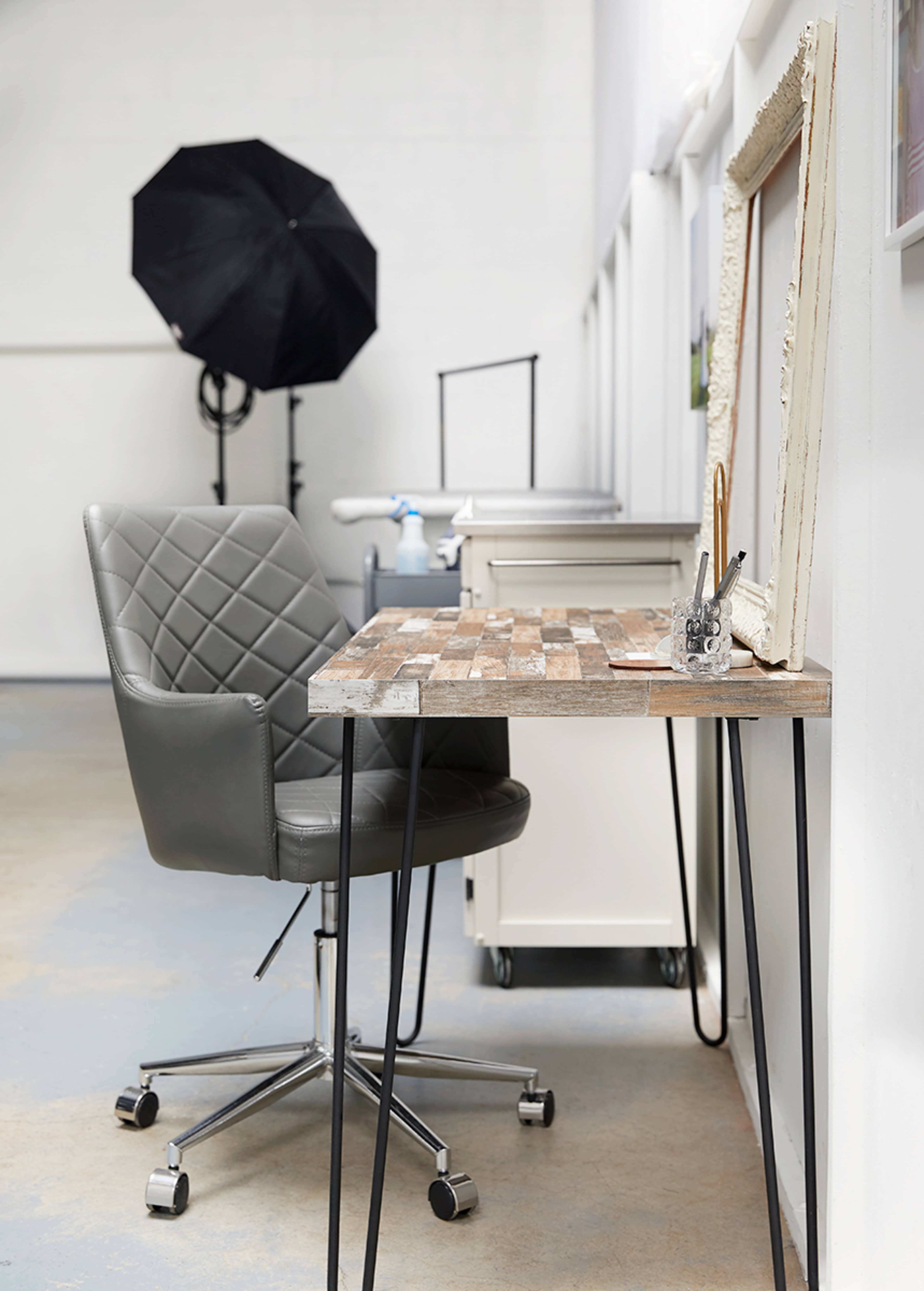 A modern workspace features a gray swivel chair next to a wooden desk with metal legs, along with a framed mirror and a photography umbrella in the background.