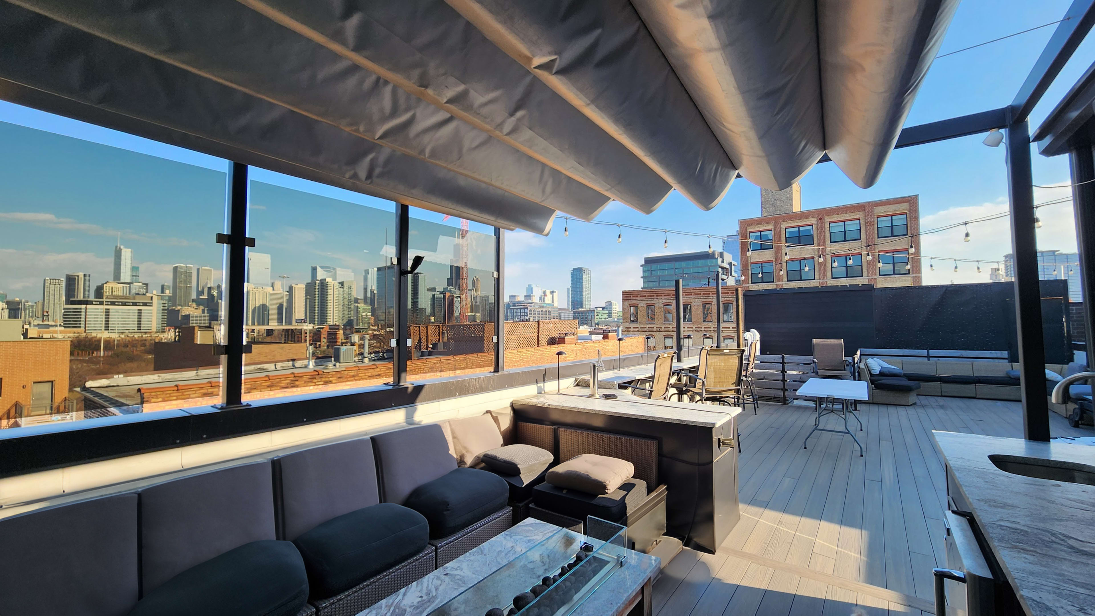 The image shows a rooftop terrace with modern seating, a glass barrier, and a view of a city skyline under a clear blue sky.