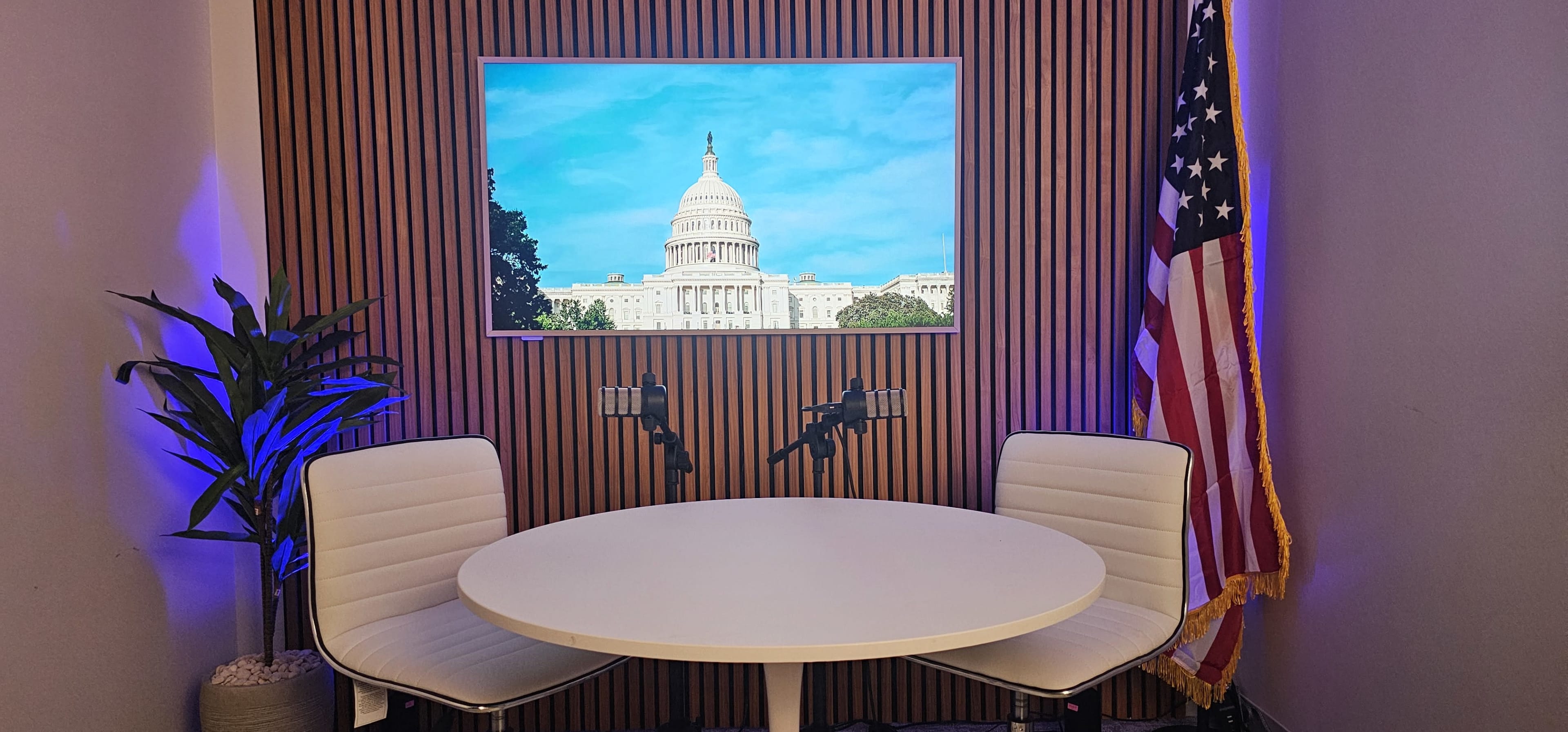 A modern meeting space features a round table with two white chairs, a backdrop of the U.S. Capitol displayed on a screen, and an American flag nearby.