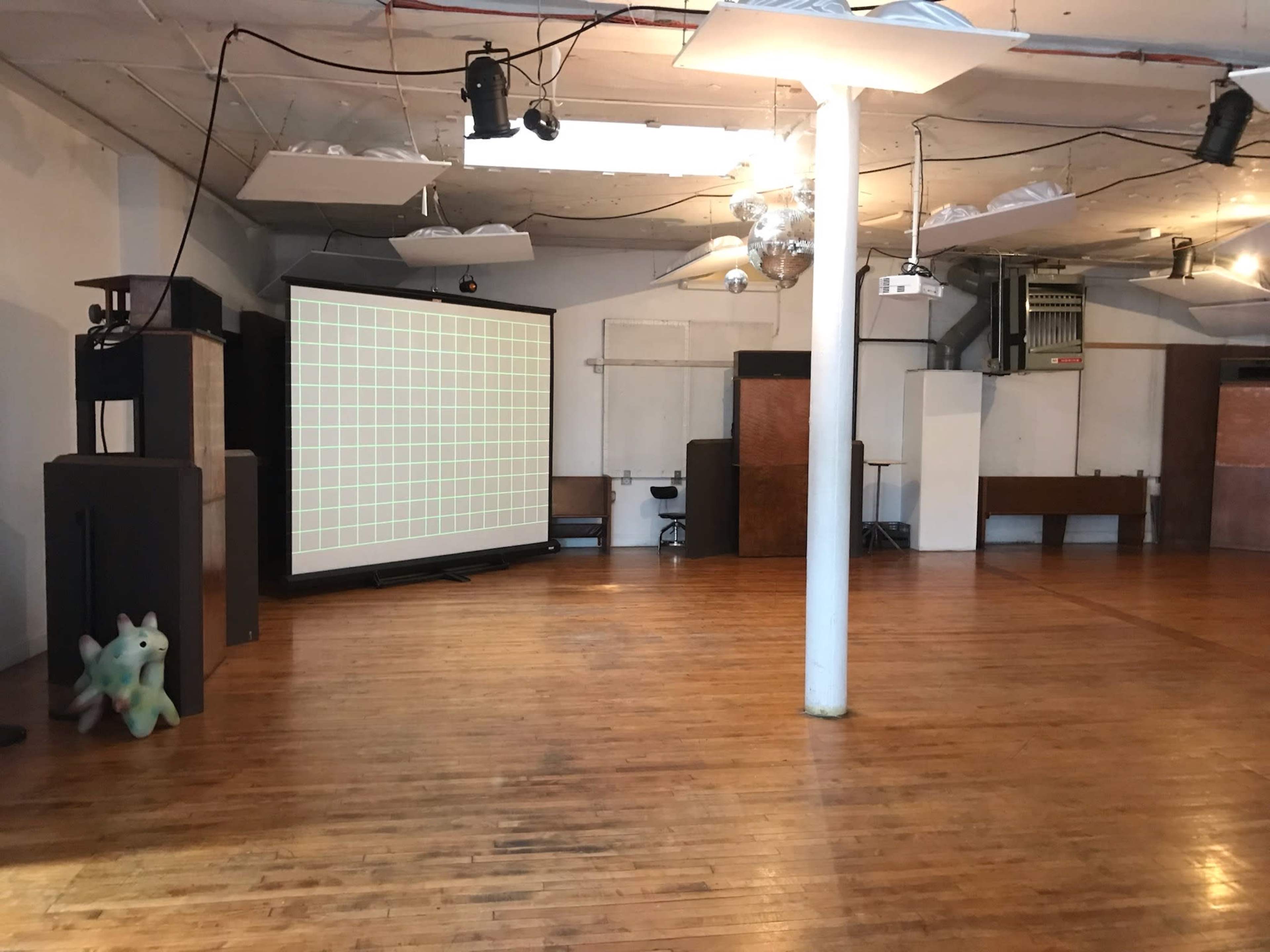 An empty indoor space with wooden flooring, featuring a large projection screen, audio equipment, and a colorful stuffed figure in the corner.