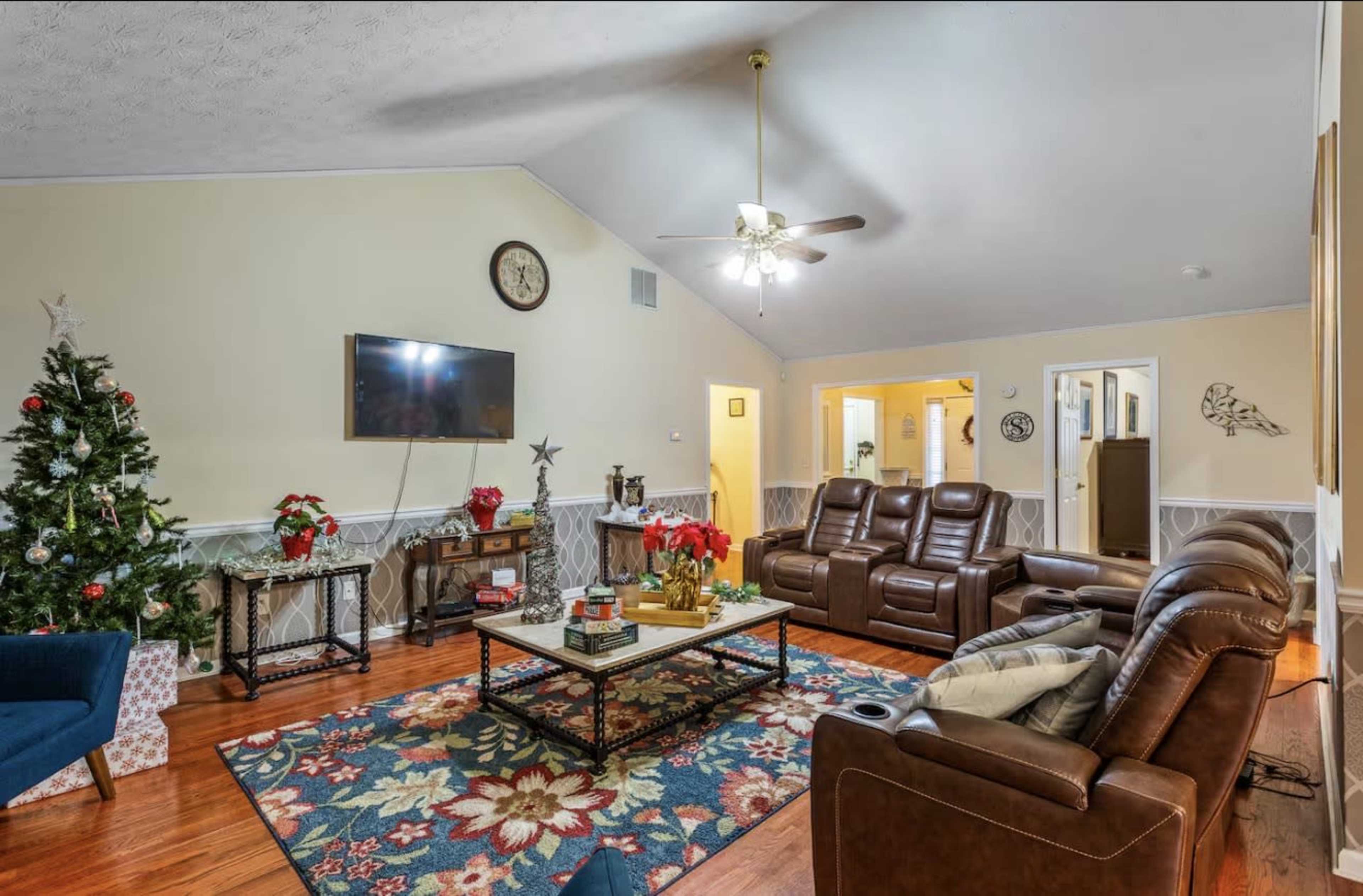 The image shows a cozy living room decorated for the holidays, featuring a Christmas tree, a large television, and comfortable seating arrangements.