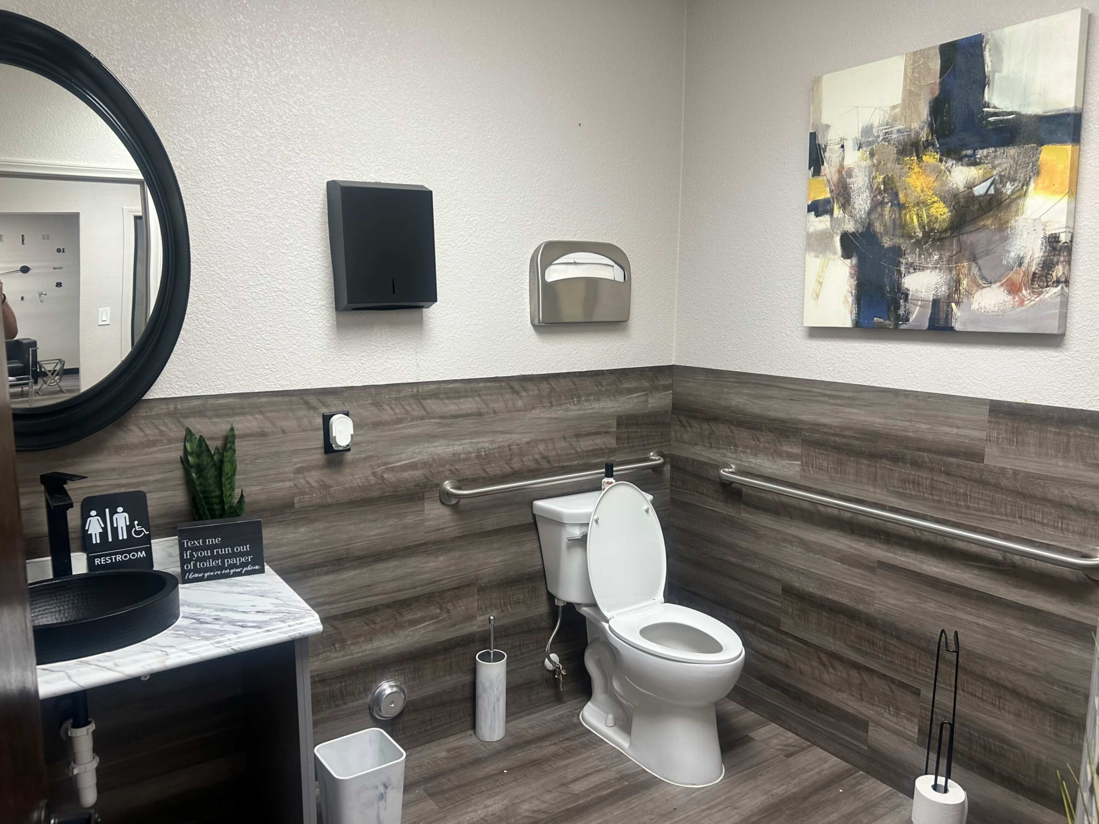 The image shows a clean bathroom featuring a toilet, a round mirror, a wall-mounted paper towel dispenser, and abstract wall art.