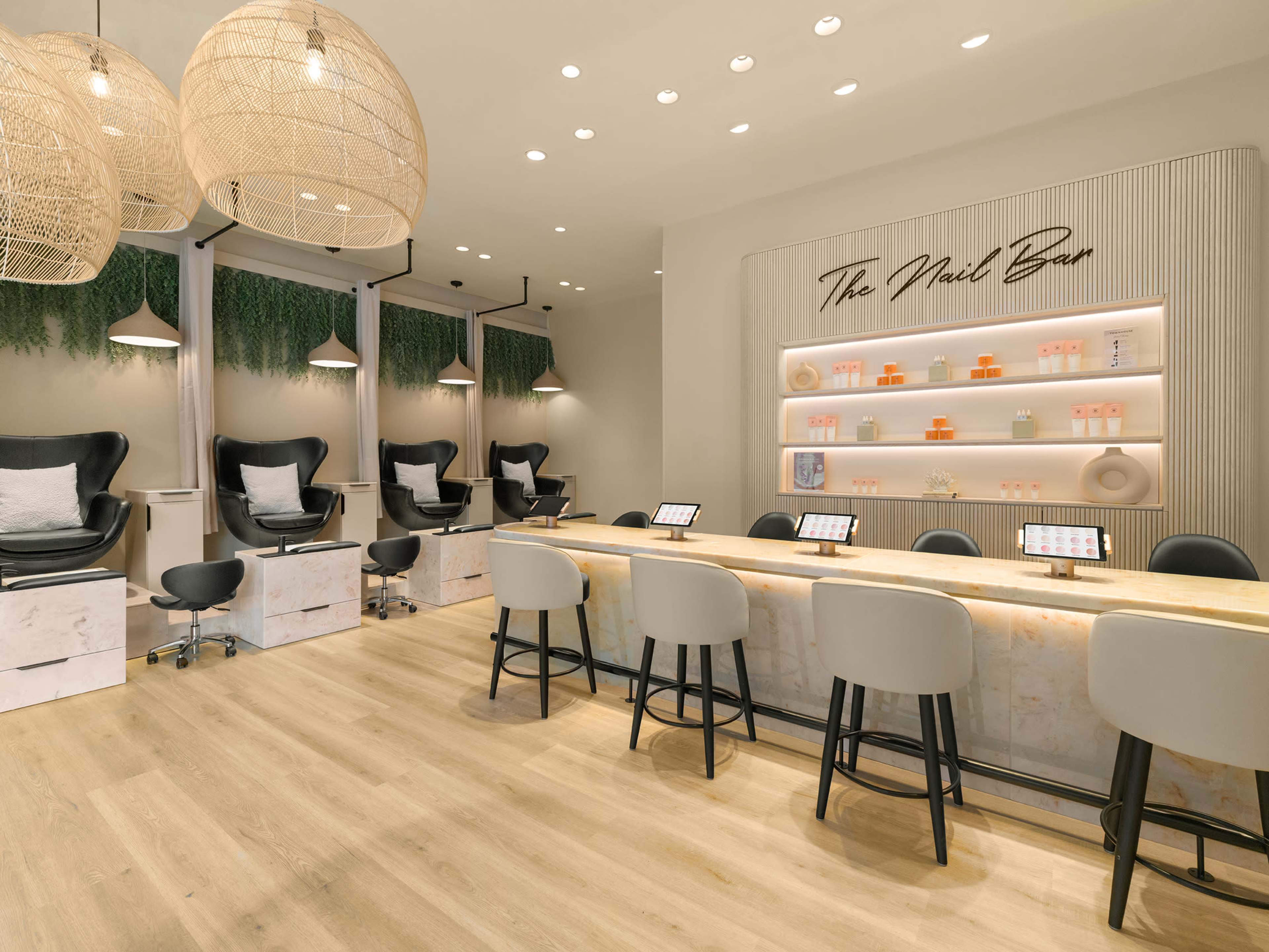 The image shows a modern nail salon with a central countertop, several pedicure stations, and stylish lighting fixtures.