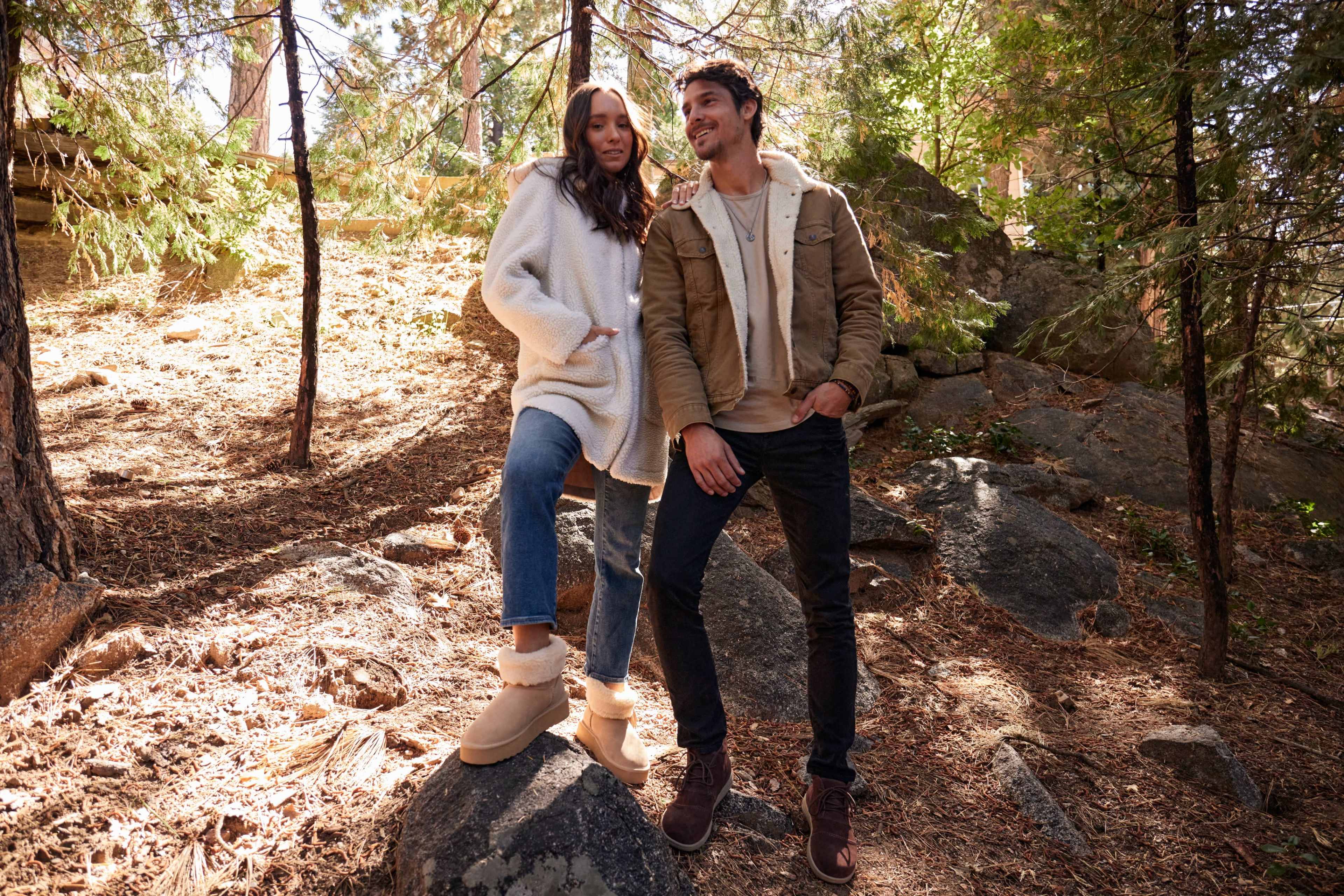 A woman in a cozy sweater and jeans stands on a rock beside a man in a tan jacket and dark pants, both posing in a wooded area with pine trees and scattered rocks.