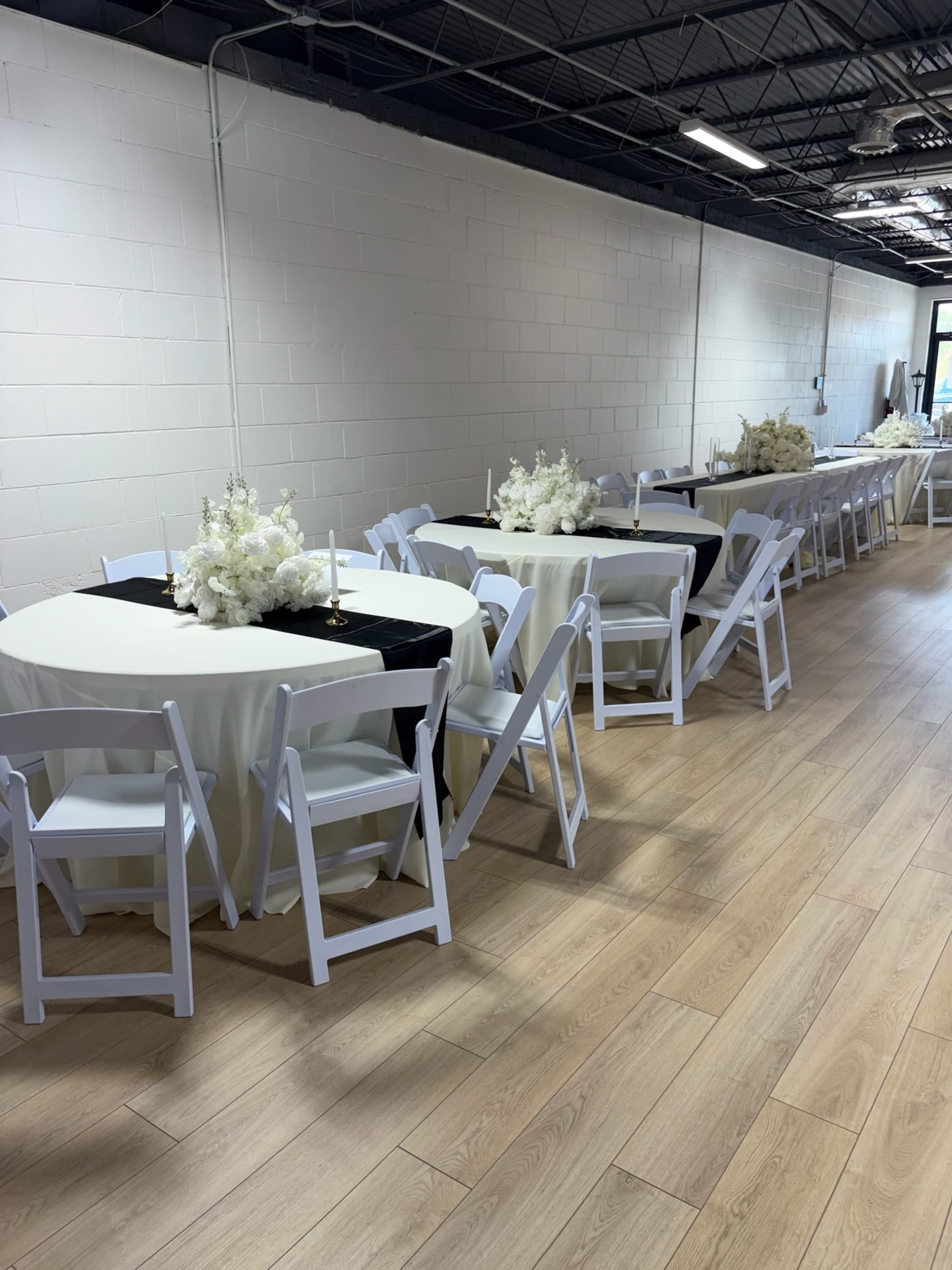 The image shows a setup of multiple white tables with black table runners, decorated with floral centerpieces, arranged in a well-lit indoor space.
