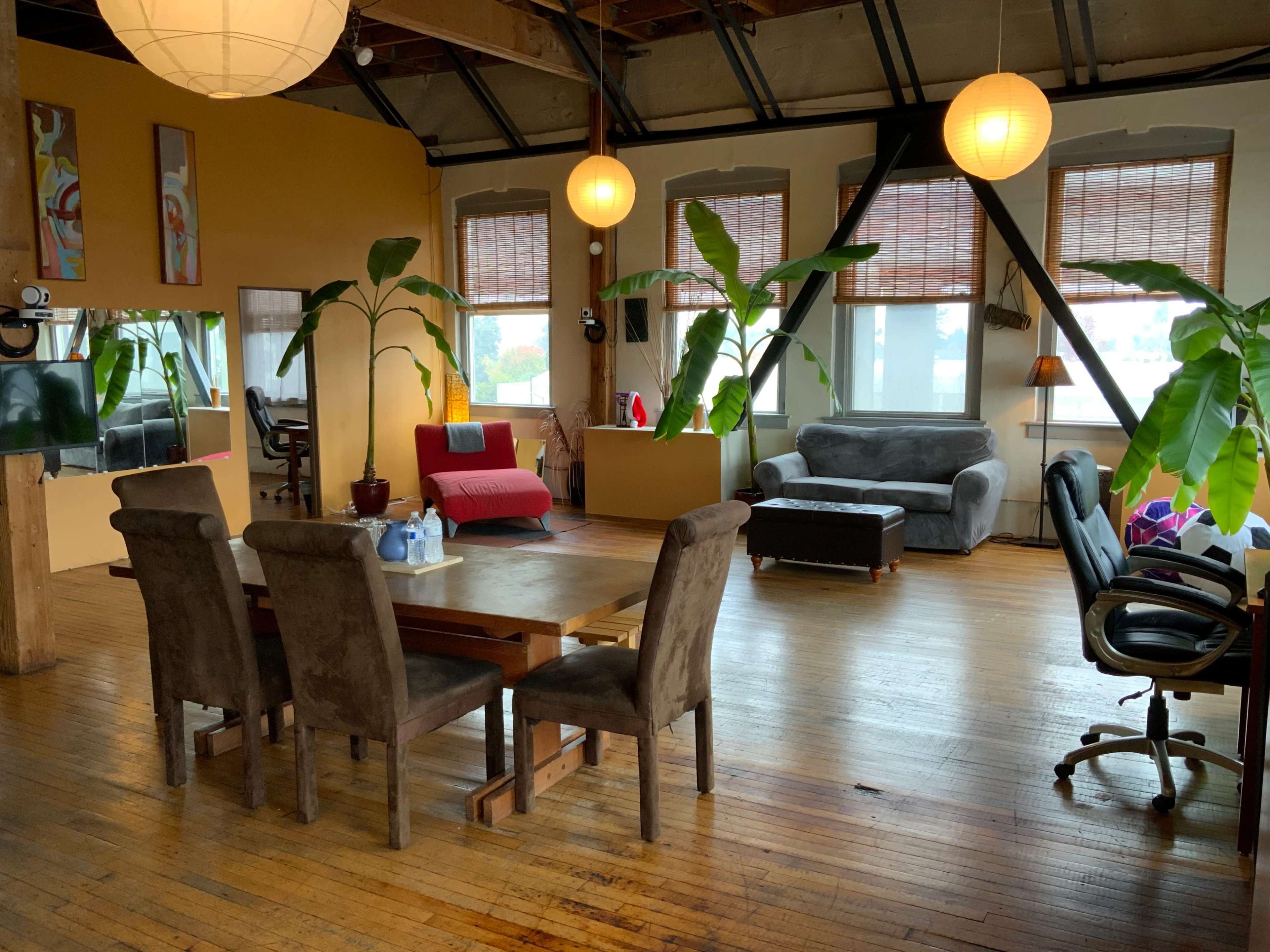 A spacious, sunlit loft with a dining area featuring a wooden table and chairs, alongside a living area with various seating options and large windows adorned with potted plants.