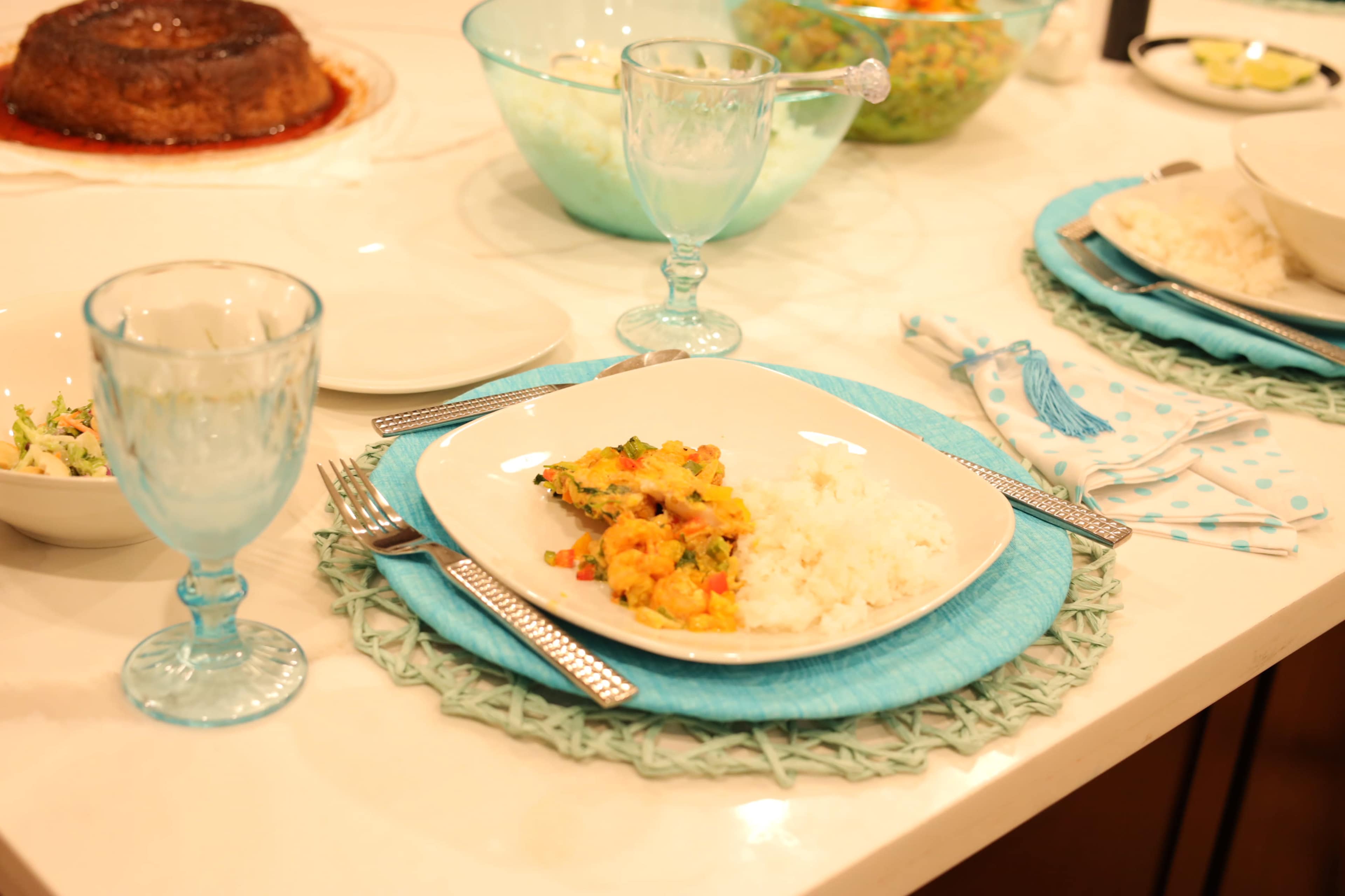 A white plate with rice and mixed vegetables is set on a blue placemat, accompanied by glassware and other dishes on a dining table.
