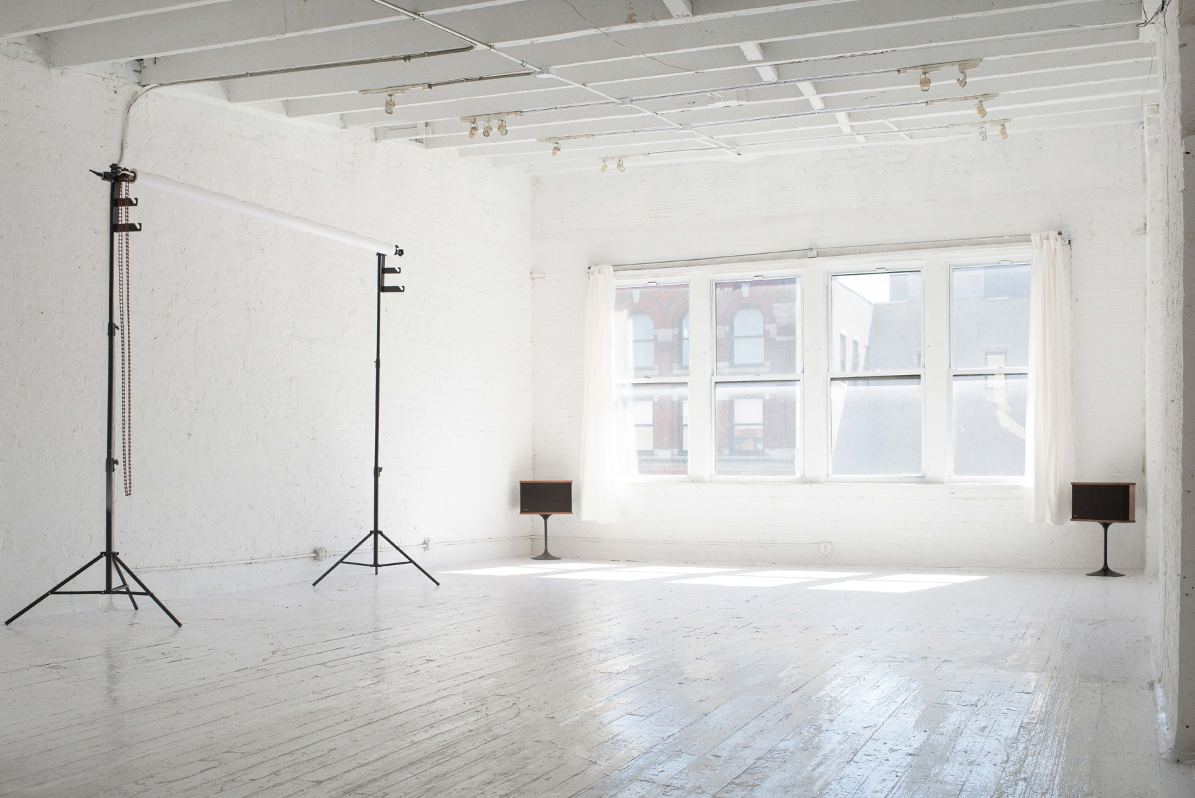 A bright, empty studio space features white walls, wooden floors, and two stands on either side, with large windows letting in natural light.