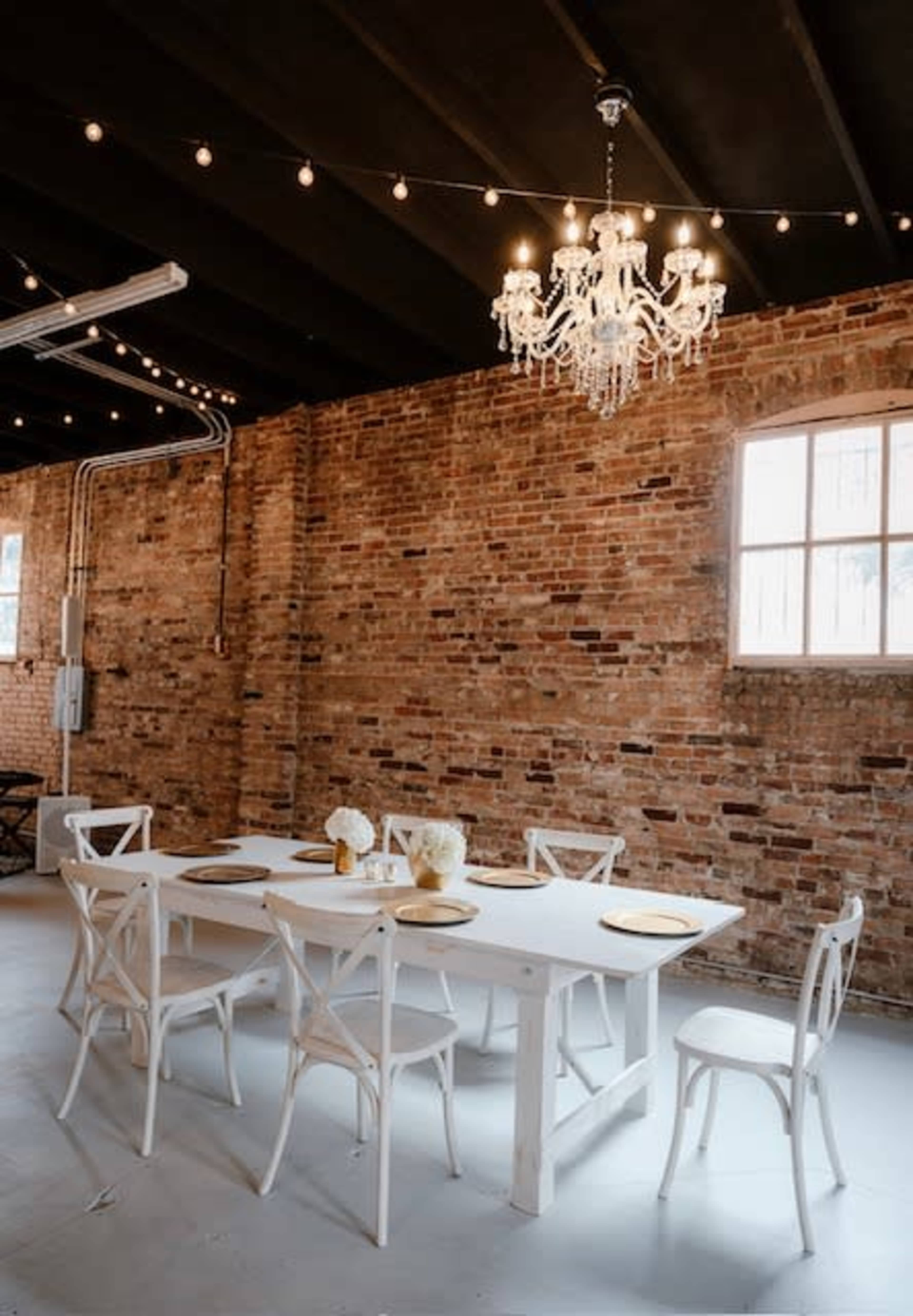 A white dining table with matching chairs is set under a chandelier in a room with exposed brick walls and string lights.