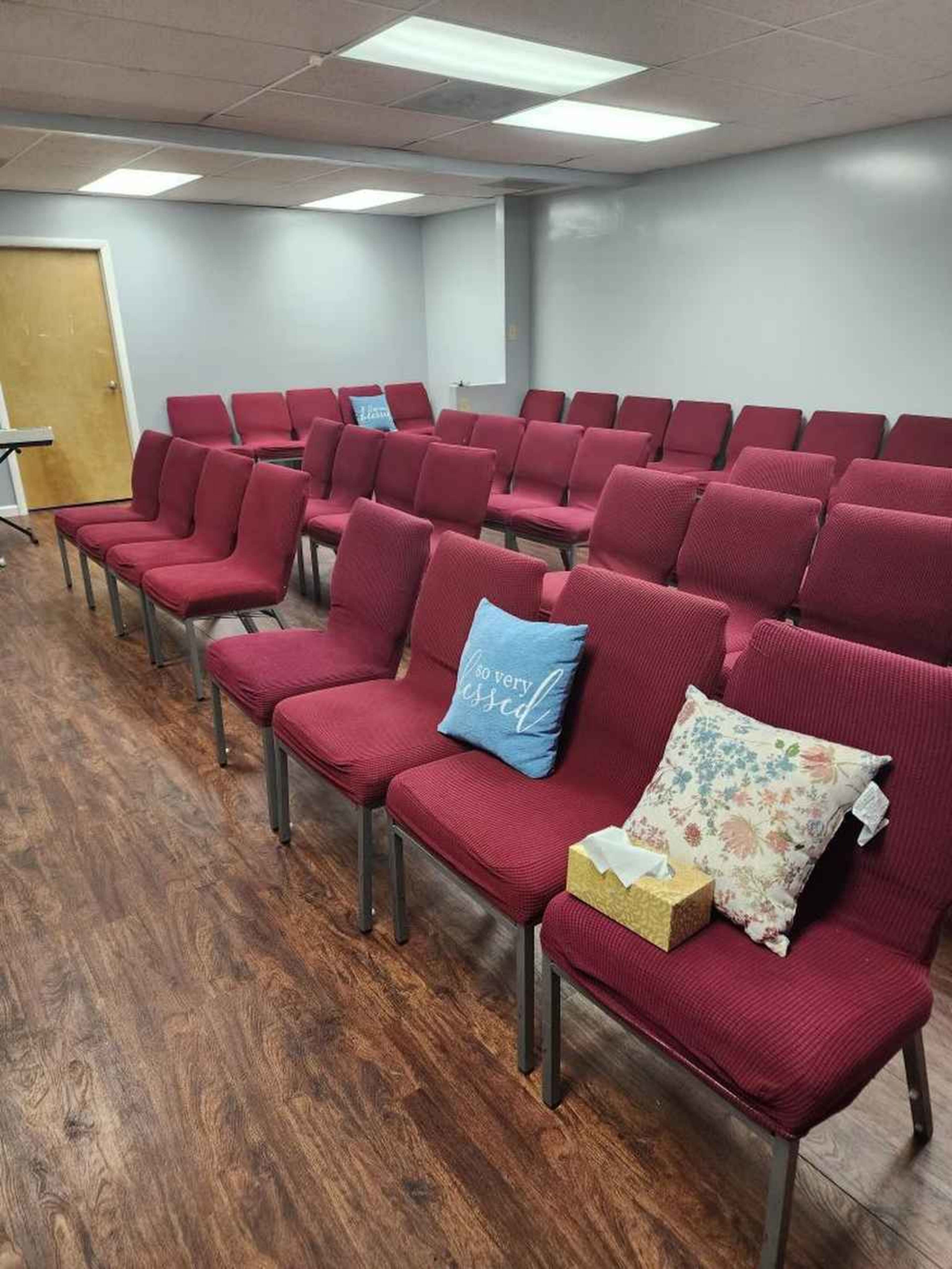 A row of empty maroon chairs arranged in a meeting room with a light wooden floor and a table in the background.