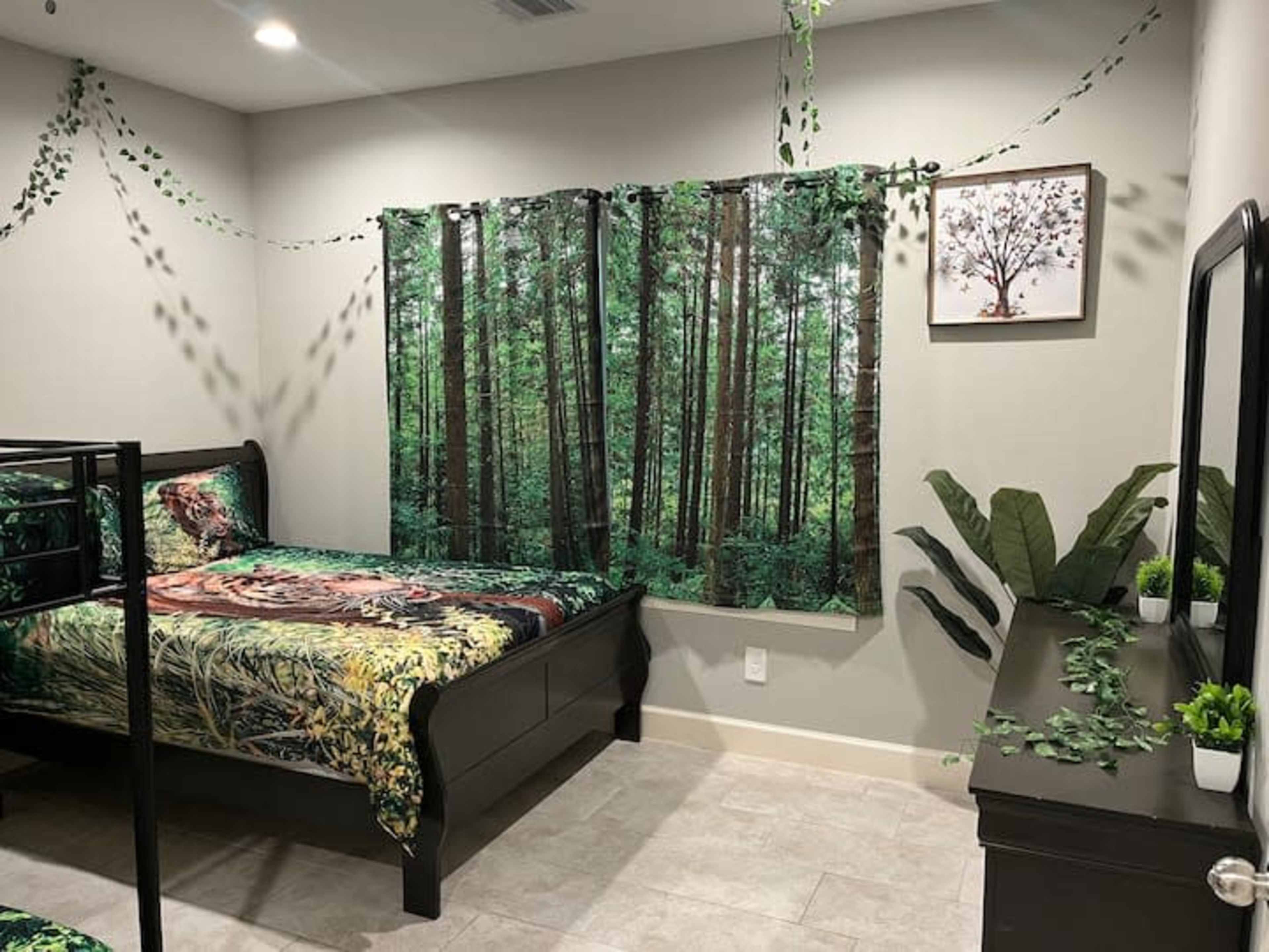 The room features a bed with a forest-themed comforter, a large window covered with nature-themed curtains, and greenery decorations on the walls and furniture.