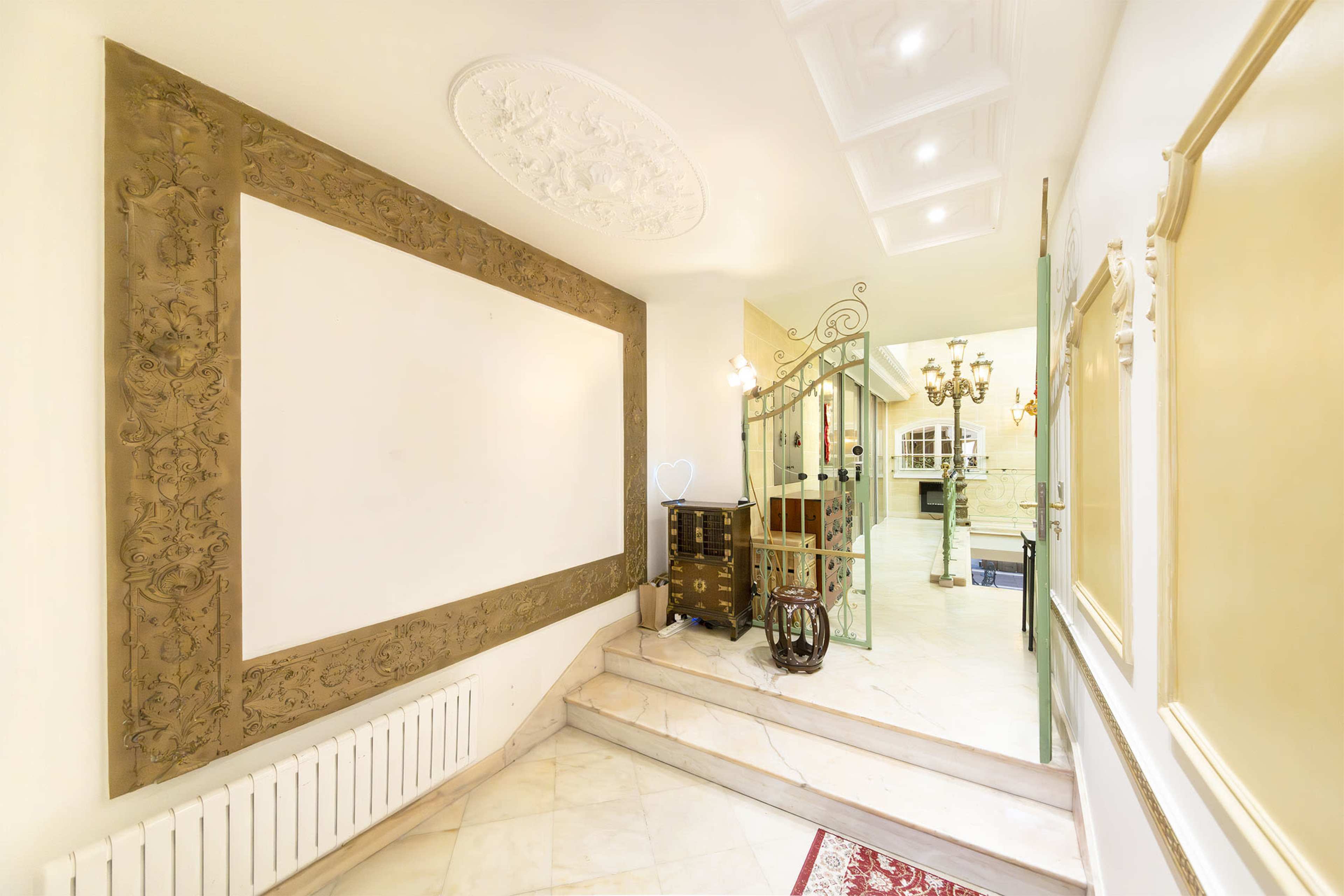 A well-lit hallway featuring decorative molding on the walls, a staircase leading to another area, and an ornate gate leading to a room with chandeliers.