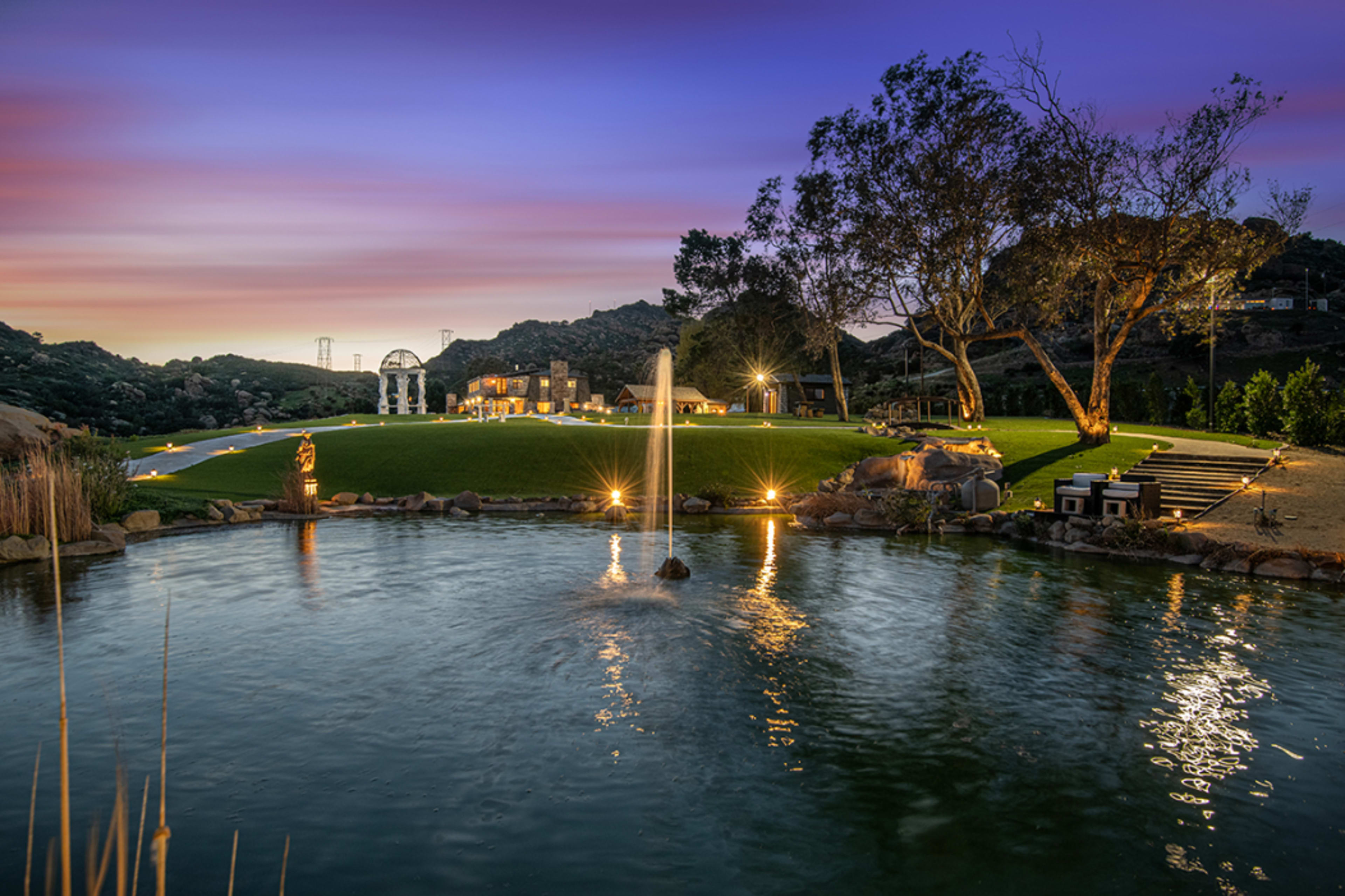 The image shows a landscaped garden with a pond, illuminated pathways, and a scenic backdrop of hills at dusk.