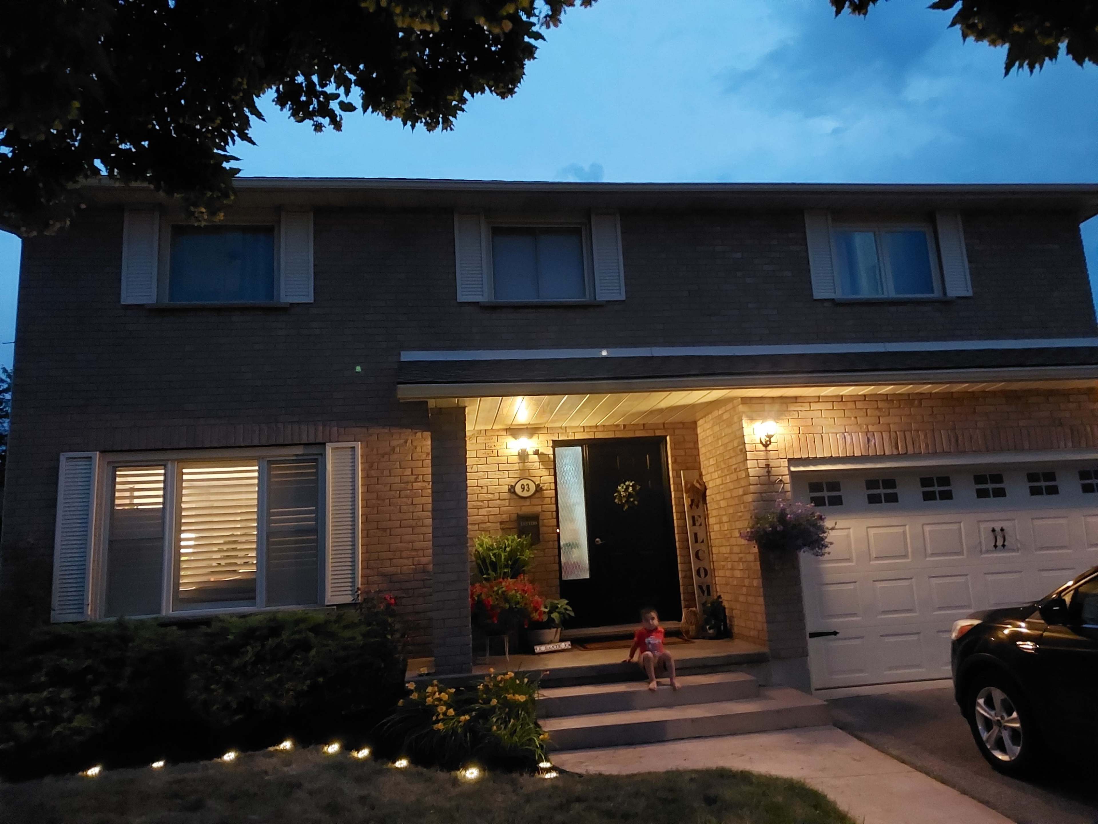 A two-story brick house with a front porch, illuminated by outdoor lighting, features a child sitting on the steps.