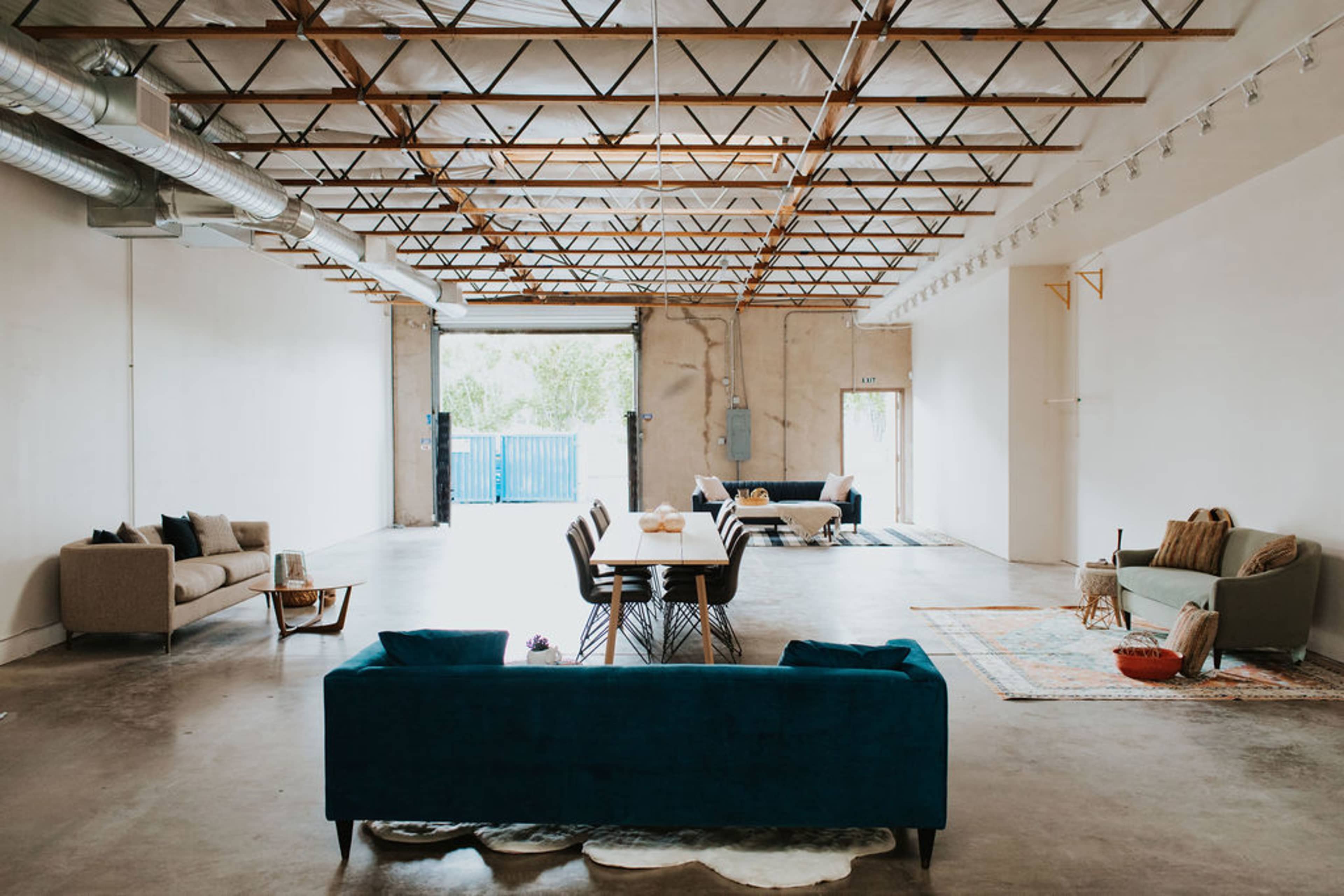 A spacious, industrial-style room features seating areas with couches and a dining table, illuminated by natural light from large doors opening to the exterior.