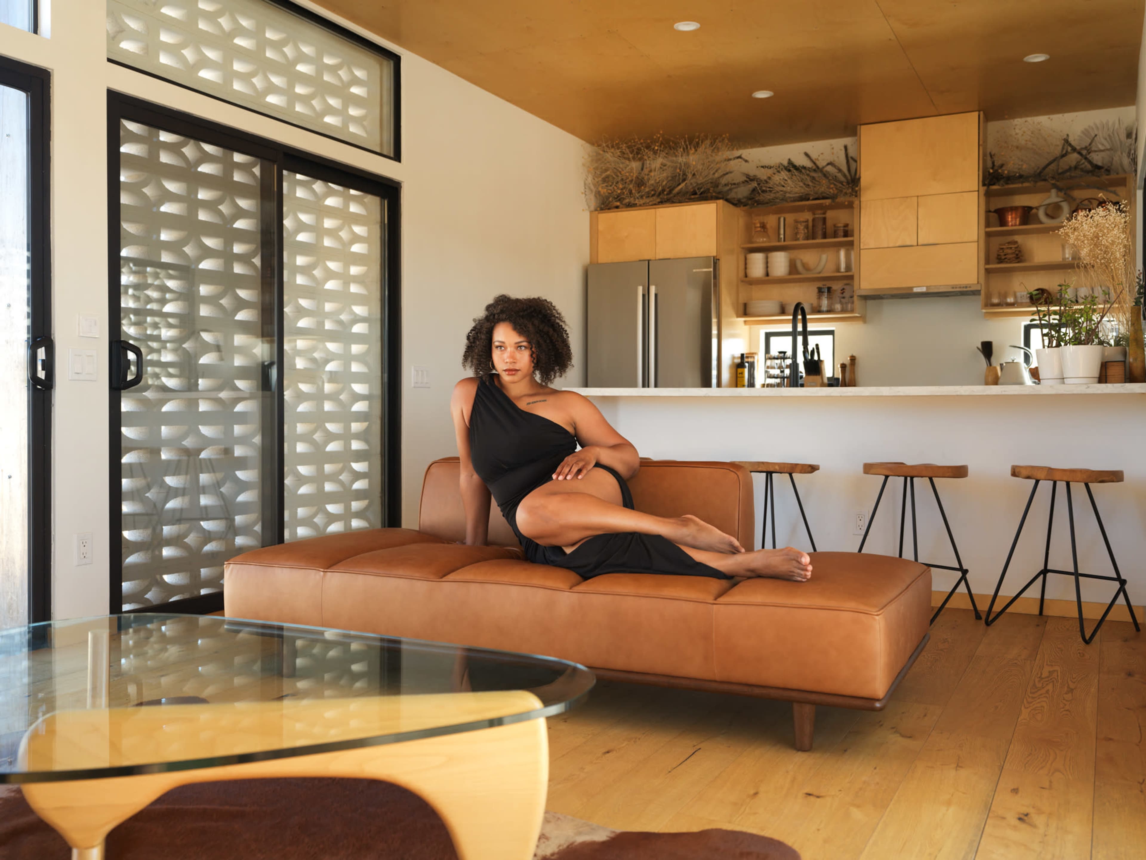 A woman sits on a brown leather chaise lounge in a modern living room, with a kitchen visible in the background.