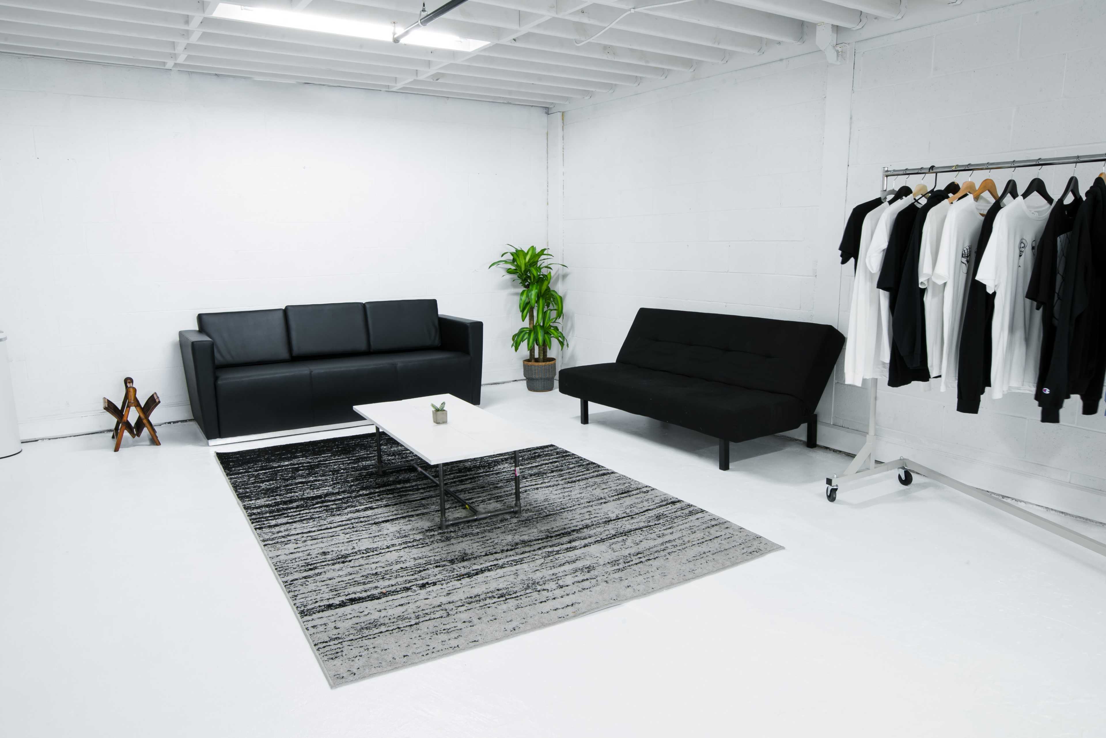 The image shows a minimalist white room featuring two sofas, a coffee table, a plant, a rug, and a clothing rack with hanging garments.