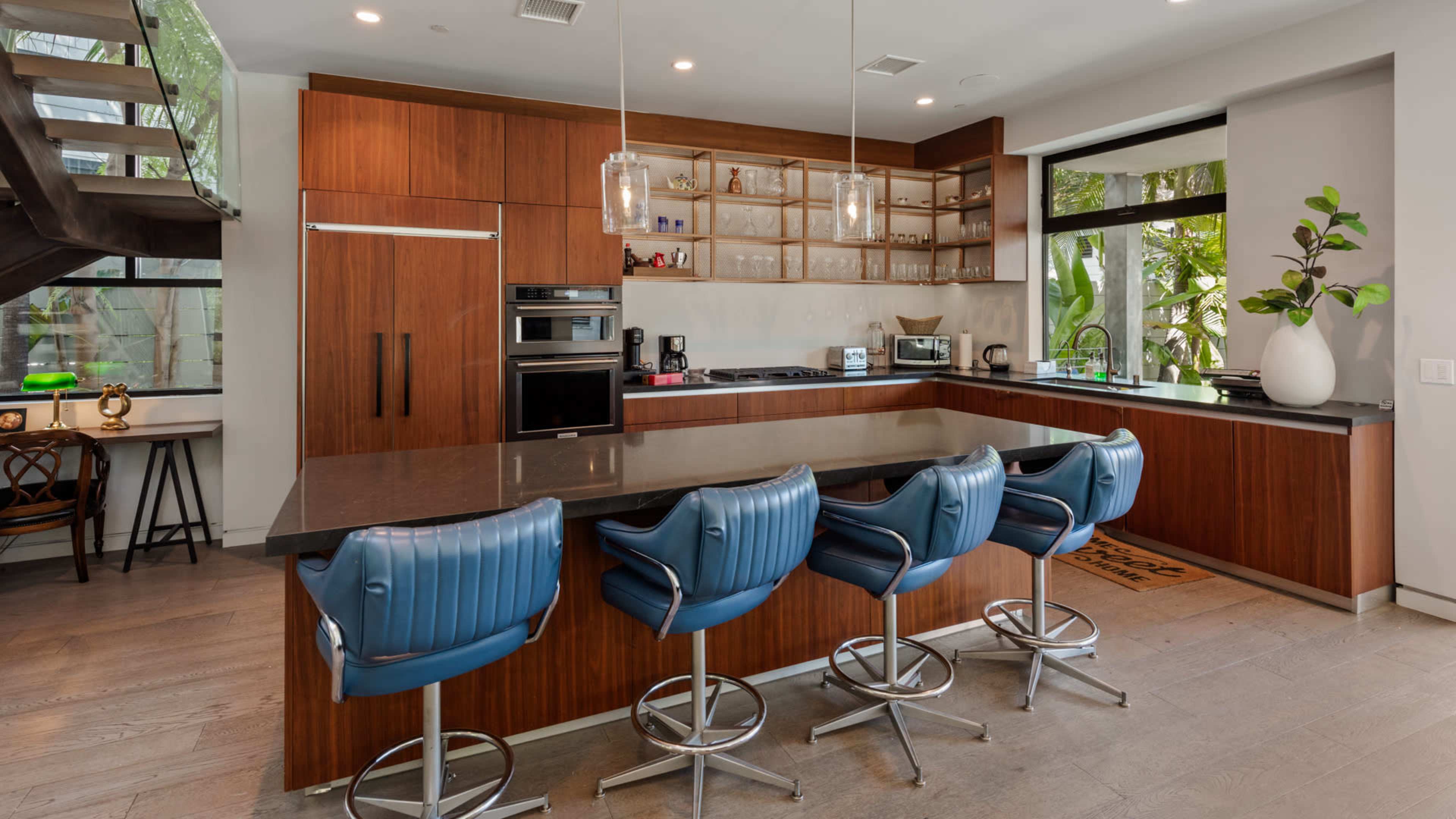 The image shows a modern kitchen with wooden cabinetry, a central island with four blue bar stools, and large windows that let in natural light.