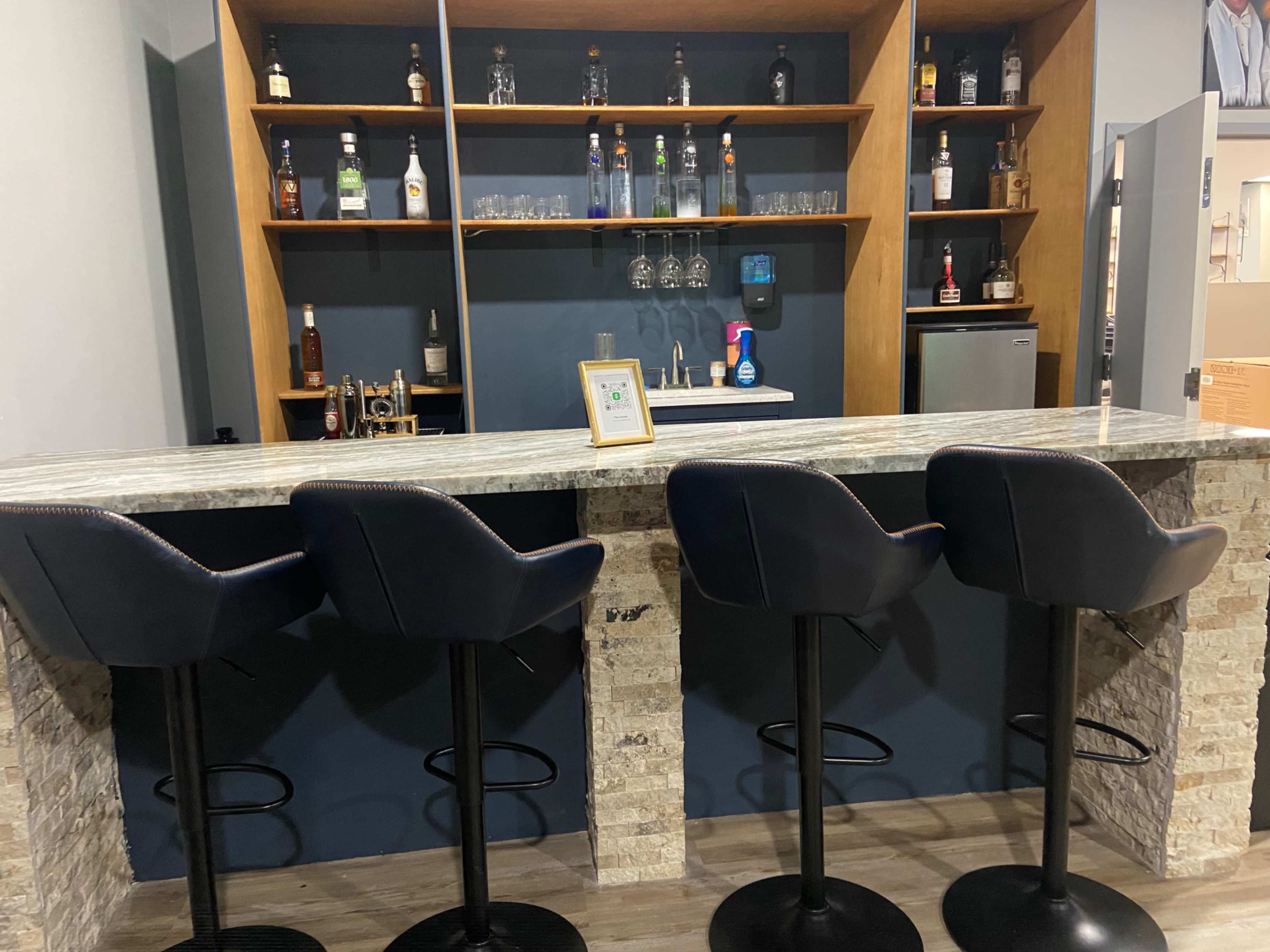 A modern bar setup features a granite counter with four black bar stools and a shelf displaying various bottles and glassware.