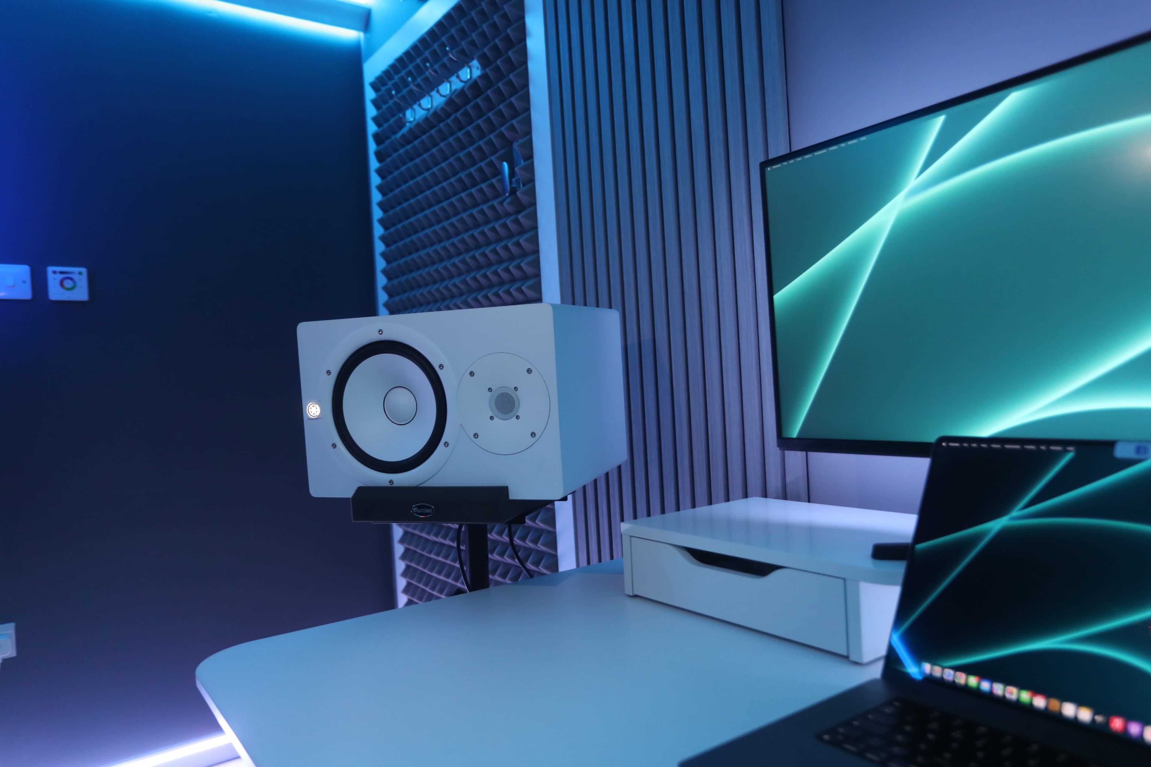 A modern workspace featuring a large monitor, a white studio monitor speaker on a stand, and a laptop on a sleek desk.