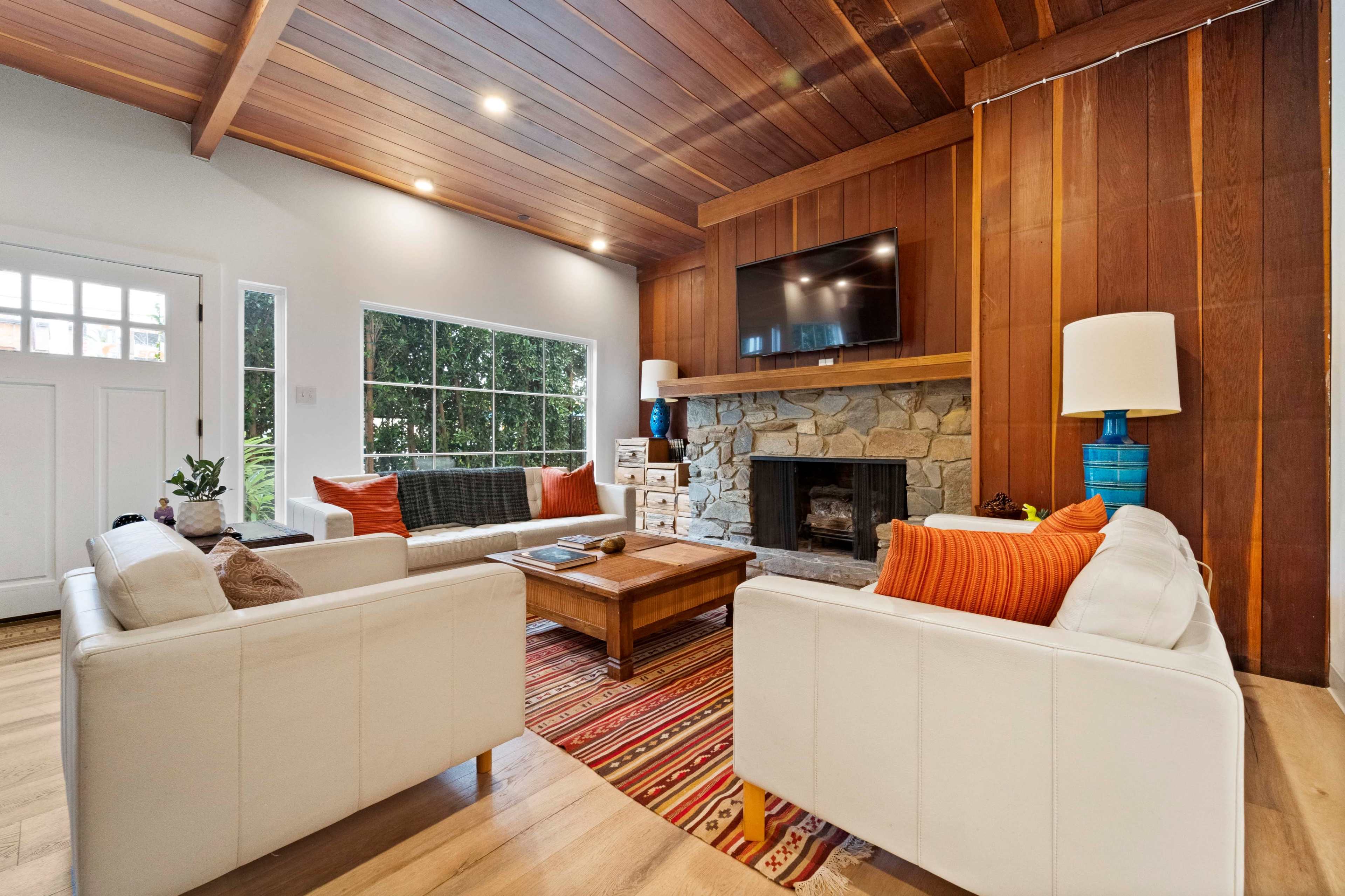 A cozy living room features a stone fireplace, wooden ceiling, and two white sofas arranged around a coffee table on a patterned rug.