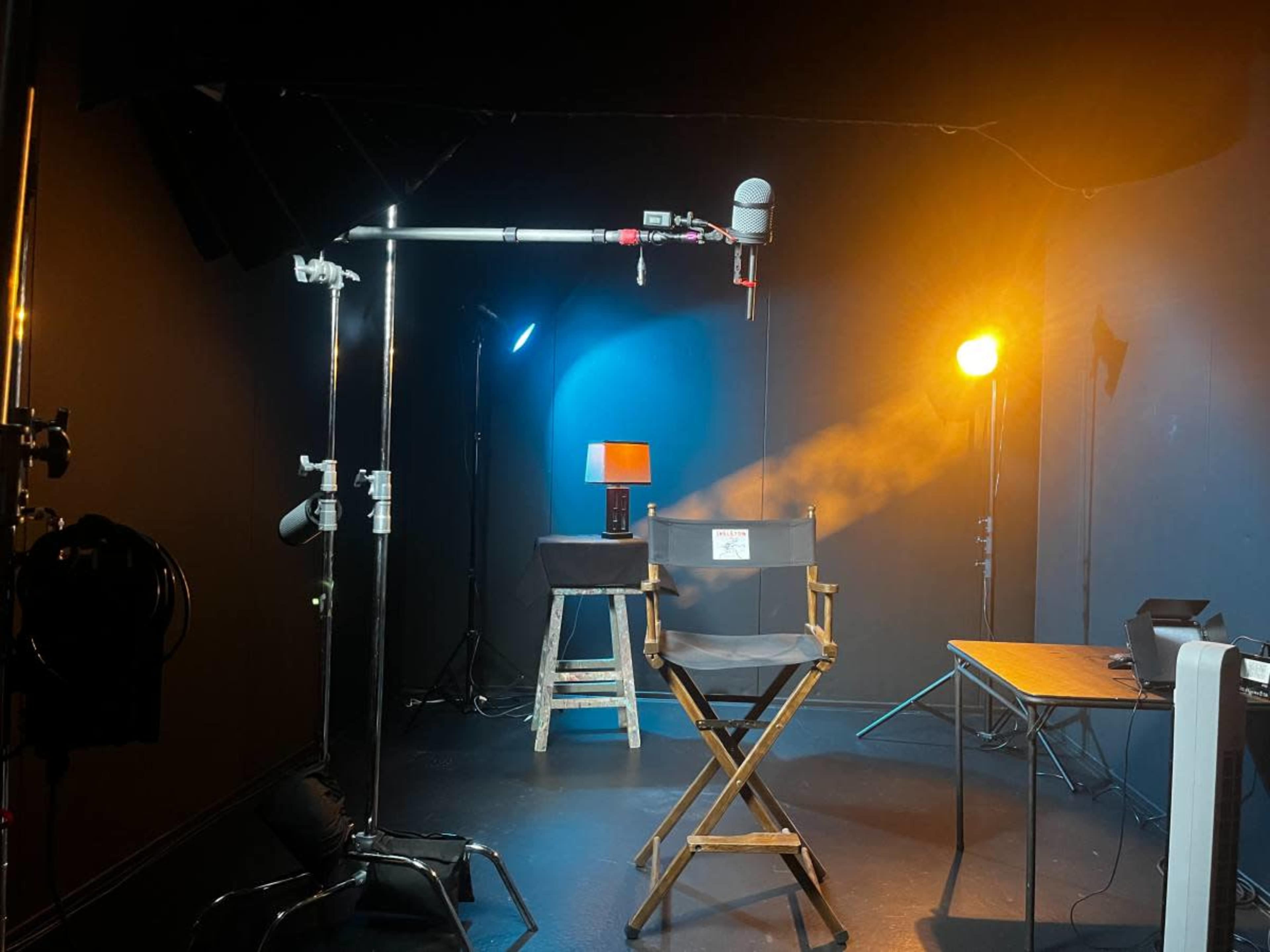 The image shows a dimly lit studio setup featuring a director's chair, a lamp on a table, and various lighting equipment.