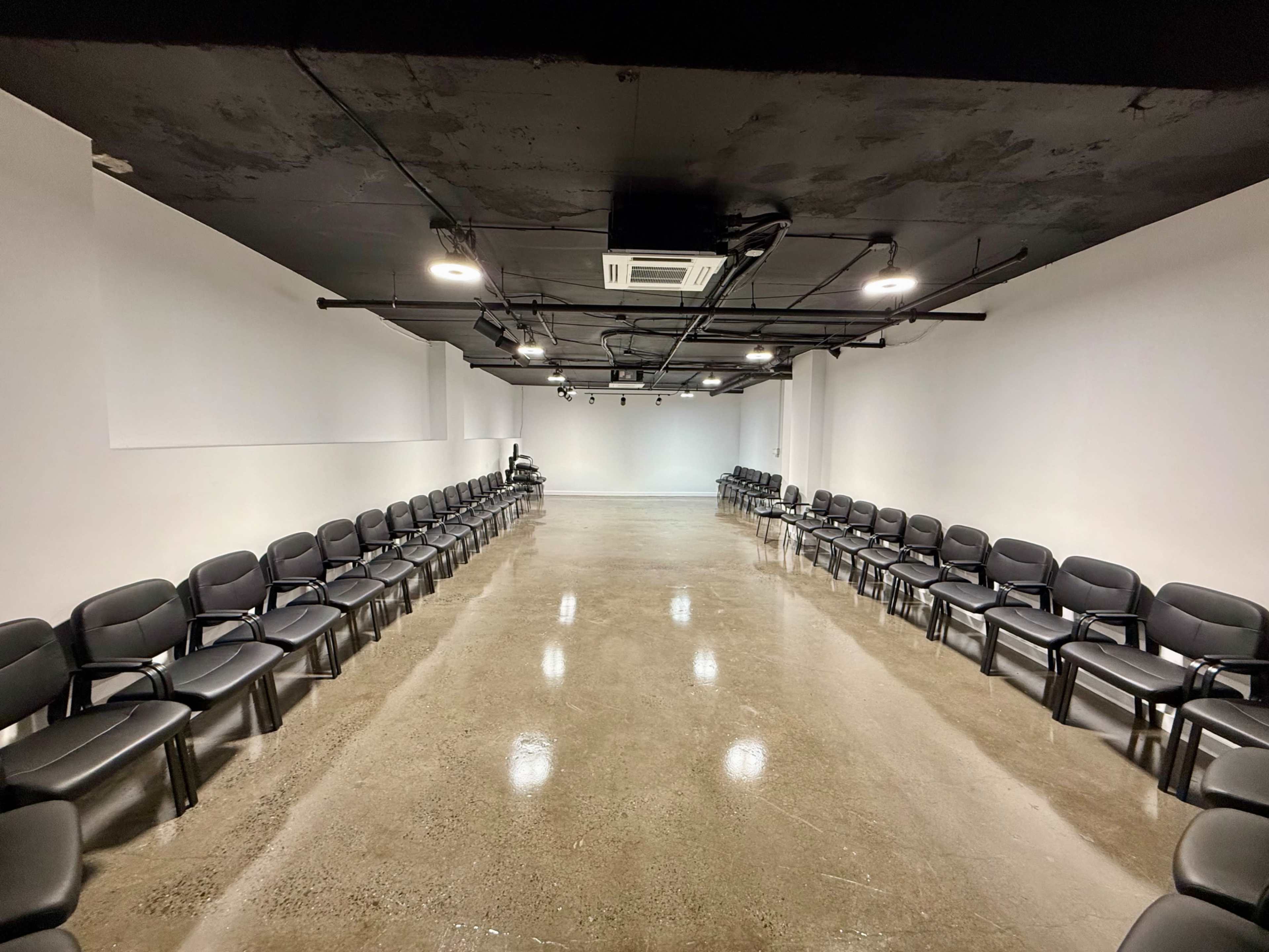 The image shows a large, empty room with two rows of black chairs arranged in a semicircle on a polished concrete floor.