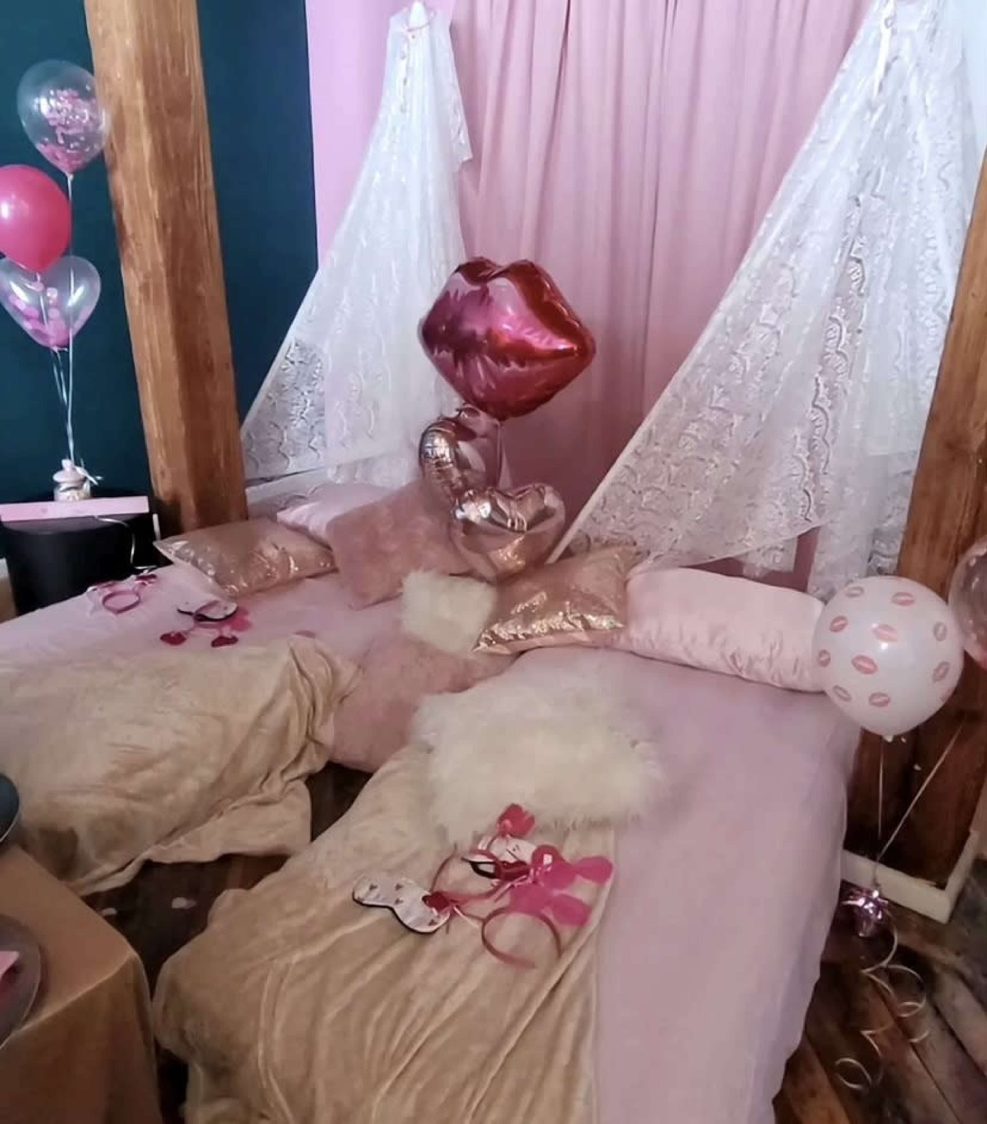 A pink-themed bedroom with two beds draped in decorative throws and pillows, surrounded by balloons and streamers.