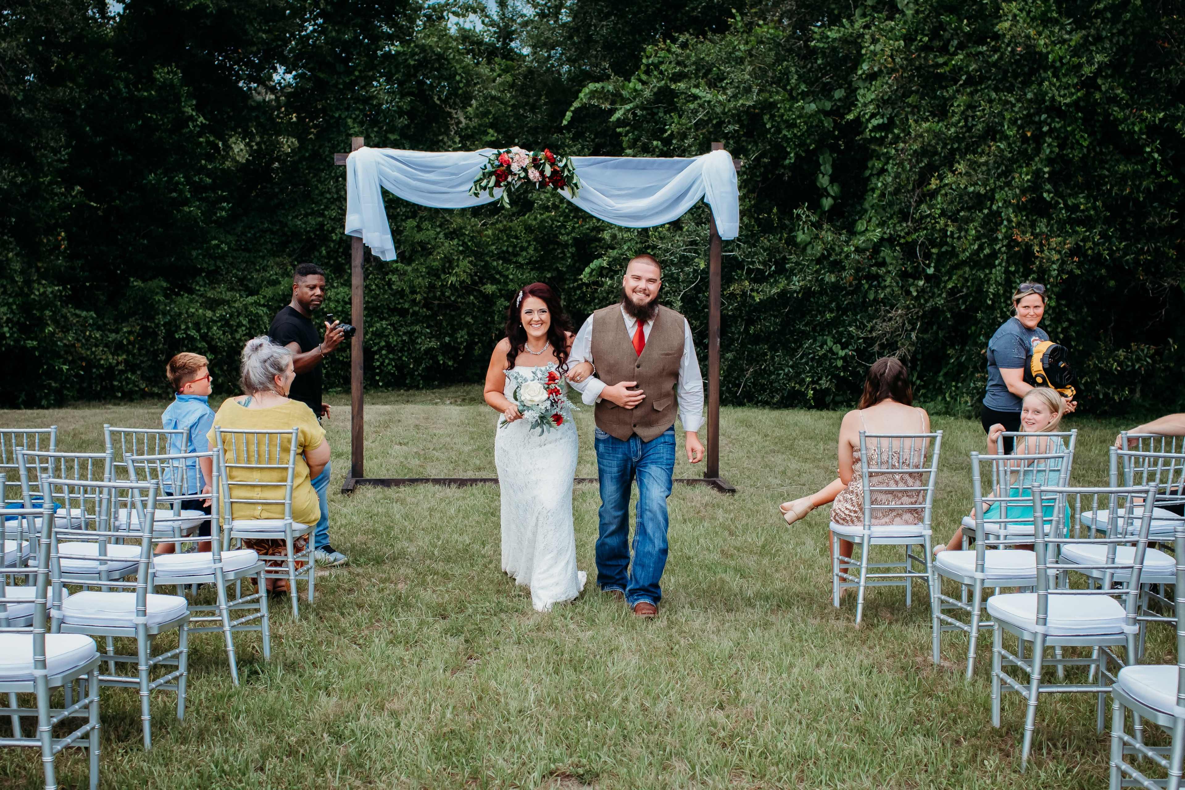 A bride and groom walk down an aisle together under an arch adorned with flowers, while seated guests look on in a grassy outdoor setting.