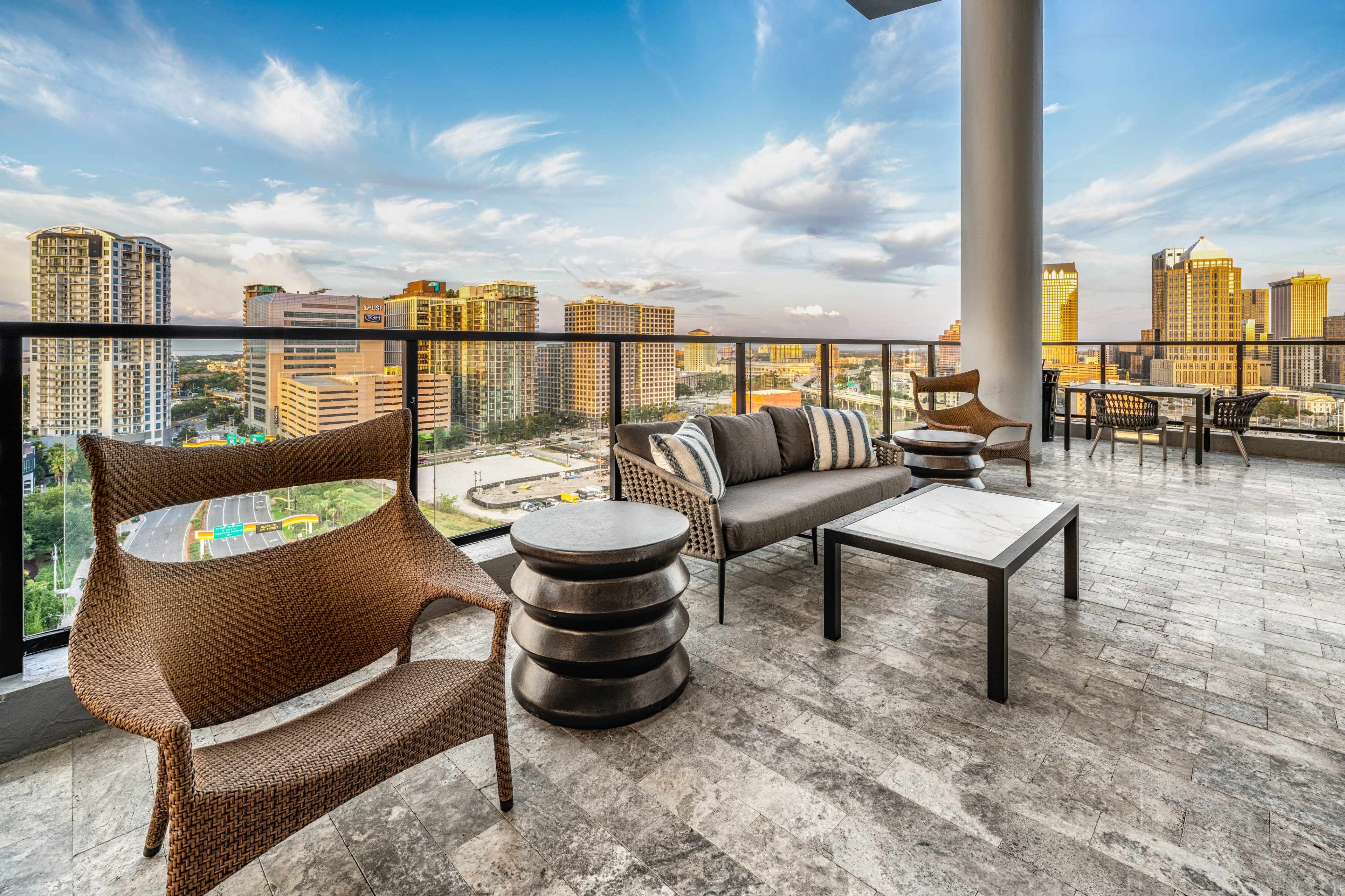 The image shows a modern rooftop patio furnished with wicker chairs and a coffee table, overlooking a city skyline under a clear blue sky.