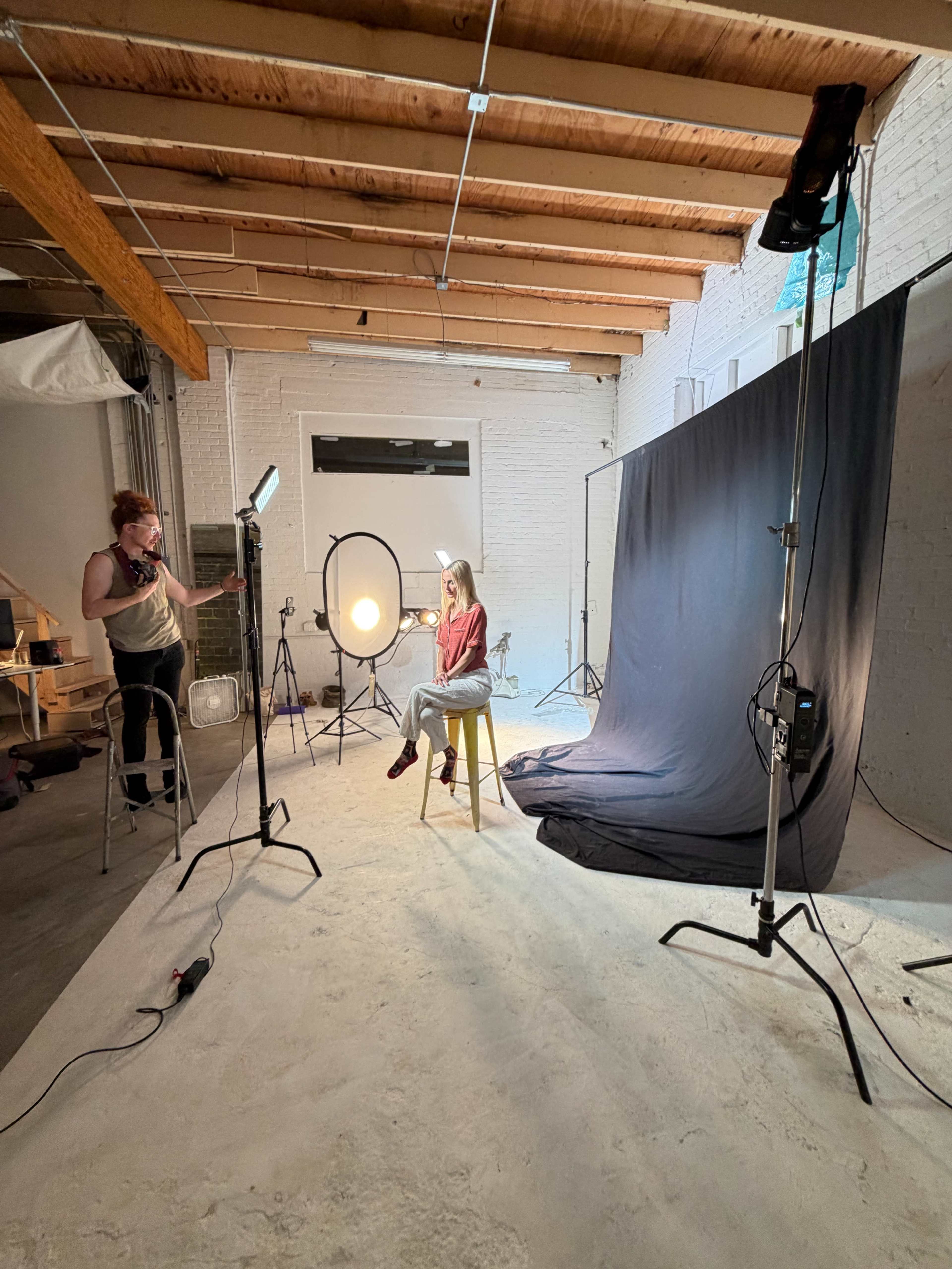 A model sits on a stool in a photography studio while a photographer adjusts lighting equipment.