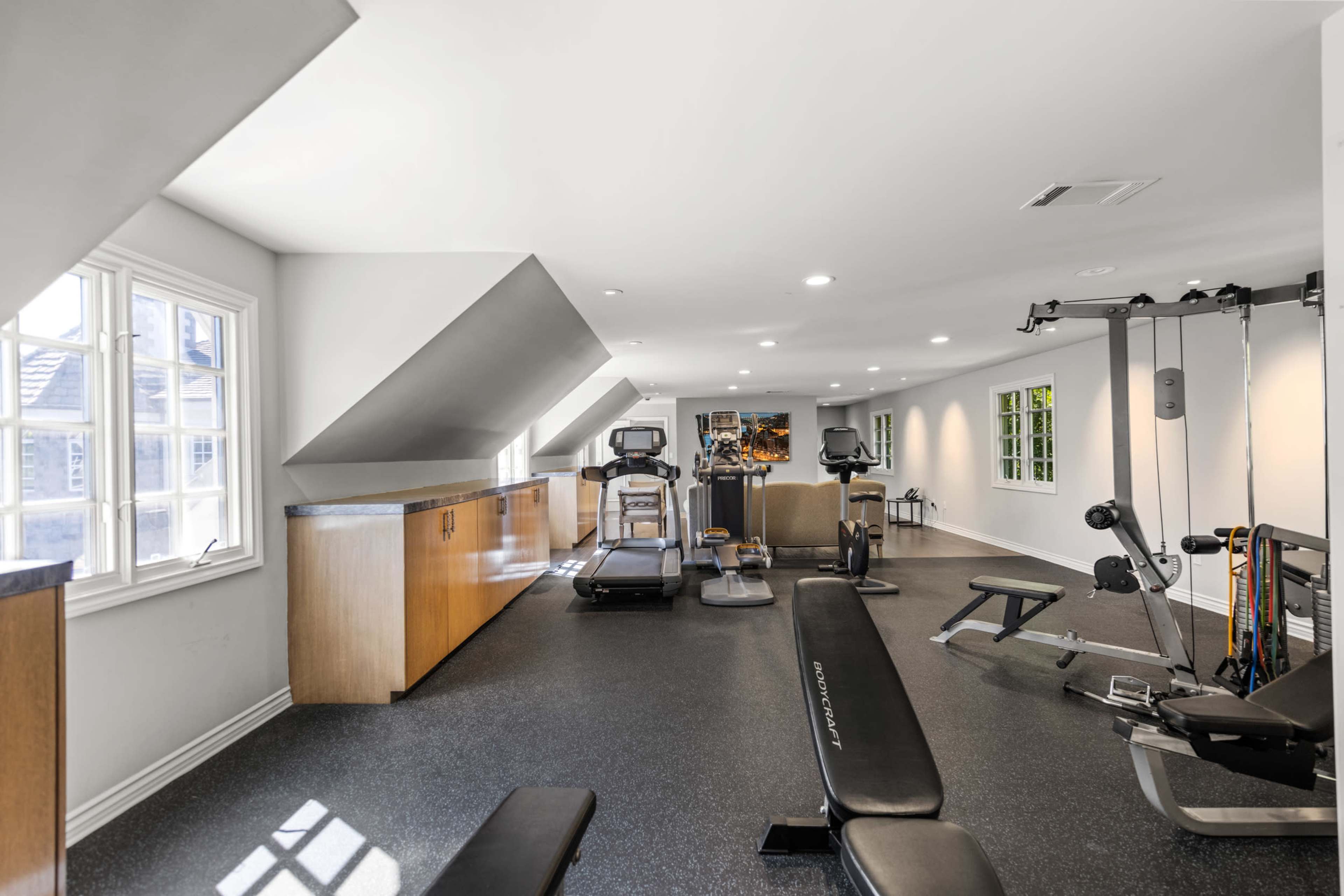 A modern home gym equipped with exercise machines and a weight bench, featuring ample natural light from multiple windows.