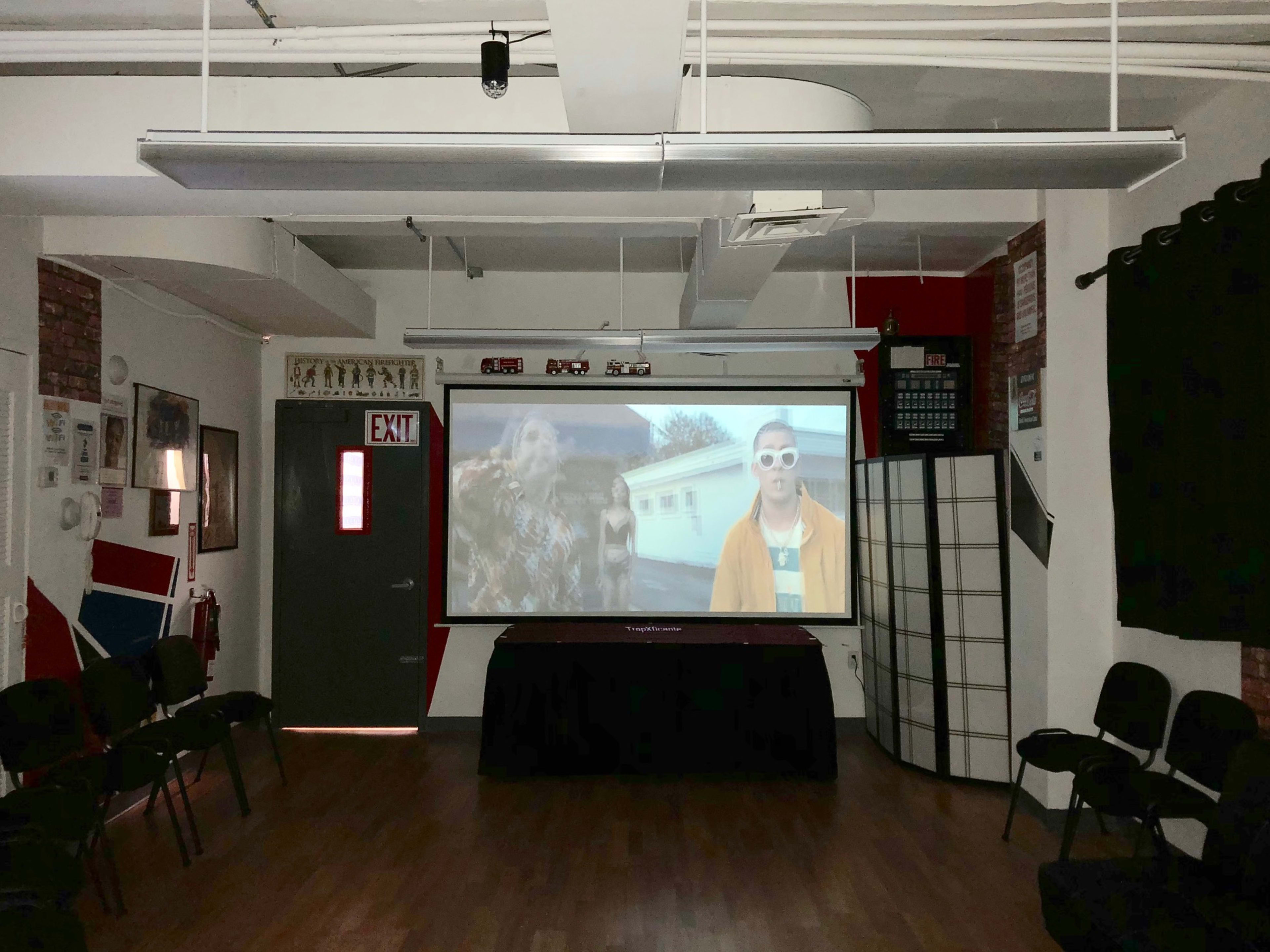 A room with chairs arranged in a semicircle faces a large screen displaying a video, with red and white walls and an exit sign visible.