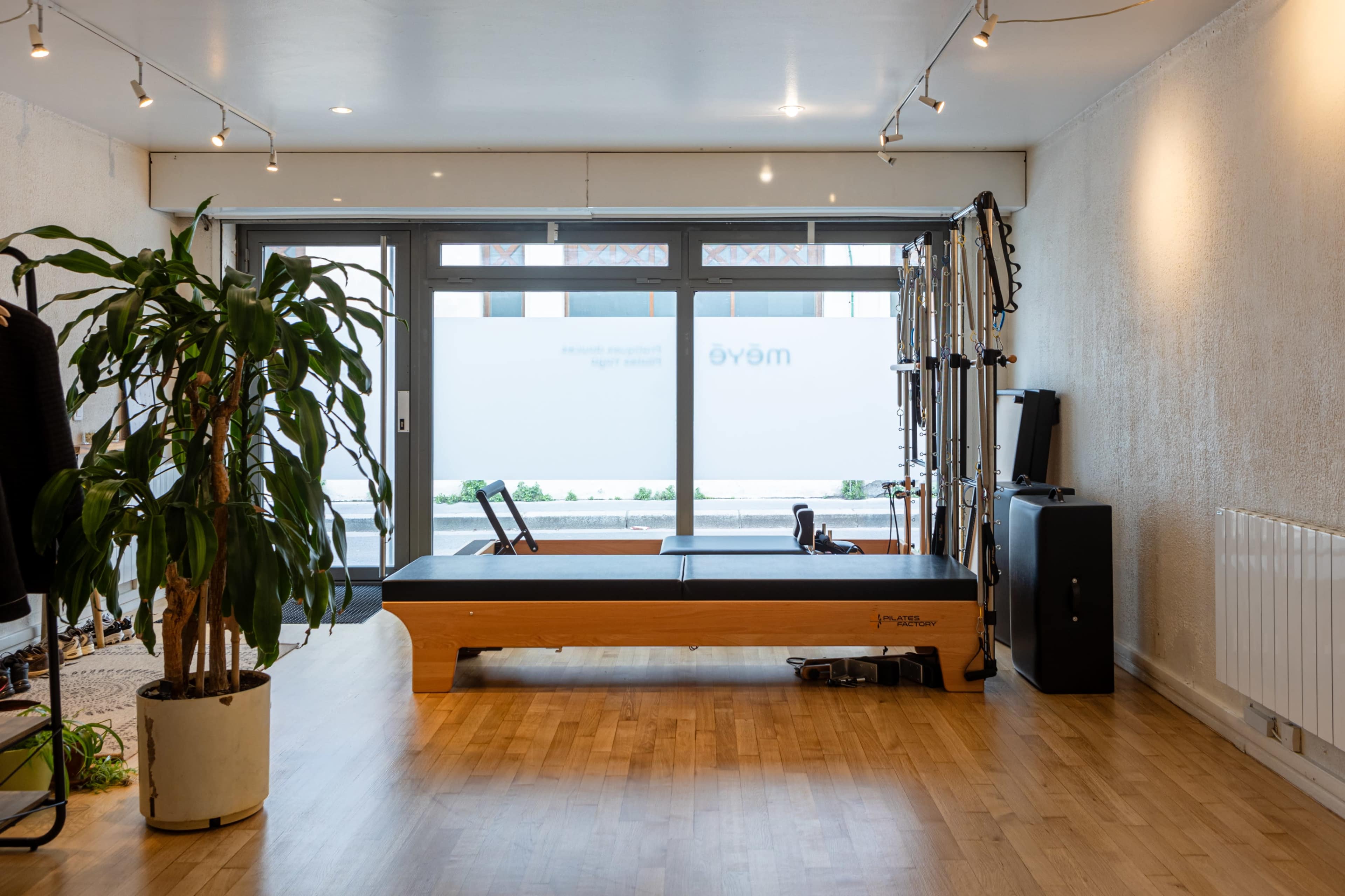 The image shows a minimalist studio space featuring a Pilates reformer, large windows allowing natural light, and a potted plant in the corner.