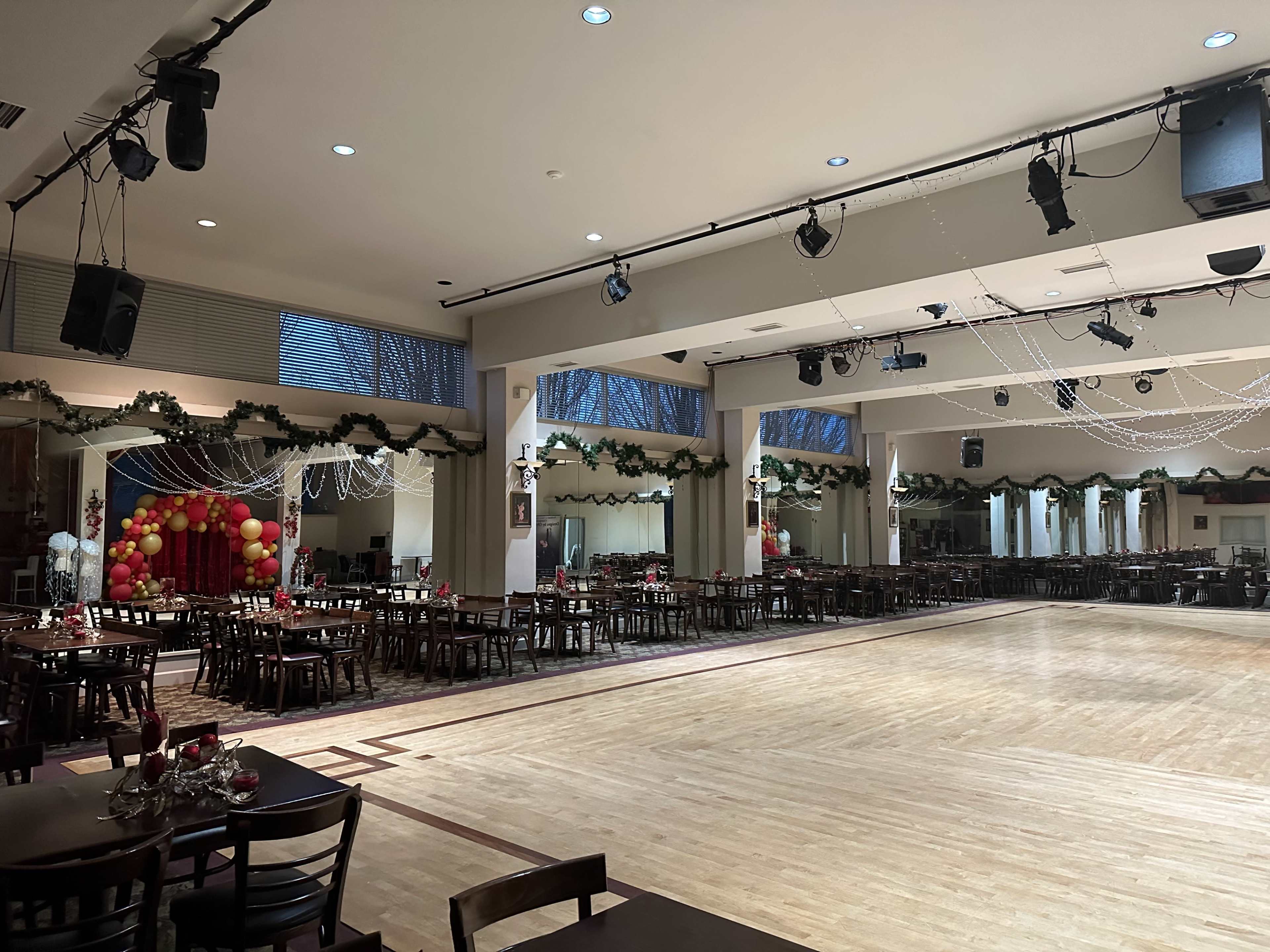 The image shows a large indoor venue set up with tables and chairs, decorated with garlands and balloon arrangements for a festive event.