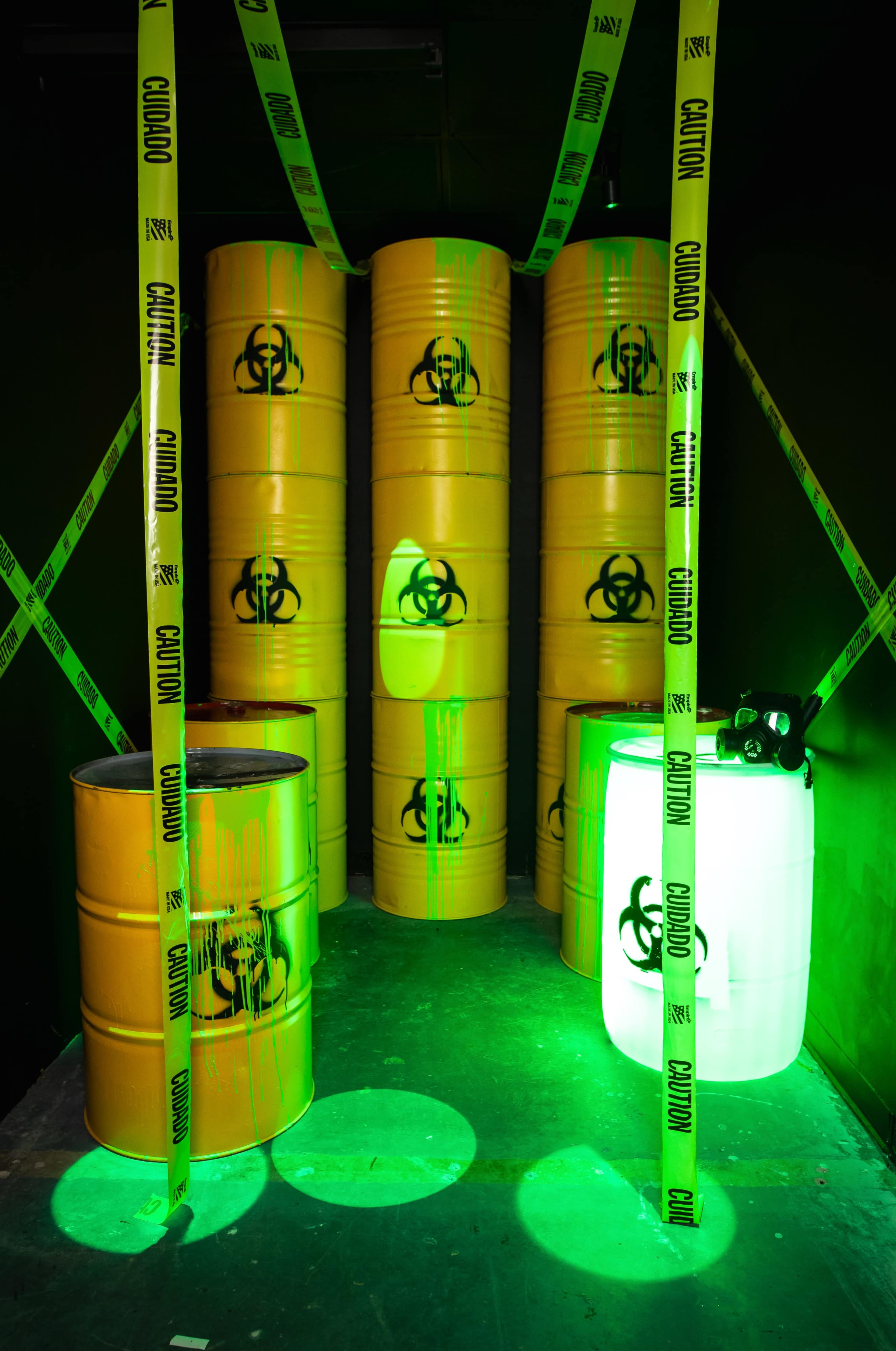 A dimly lit space with biohazard-marked yellow barrels, surrounded by caution tape and illuminated by green lighting.