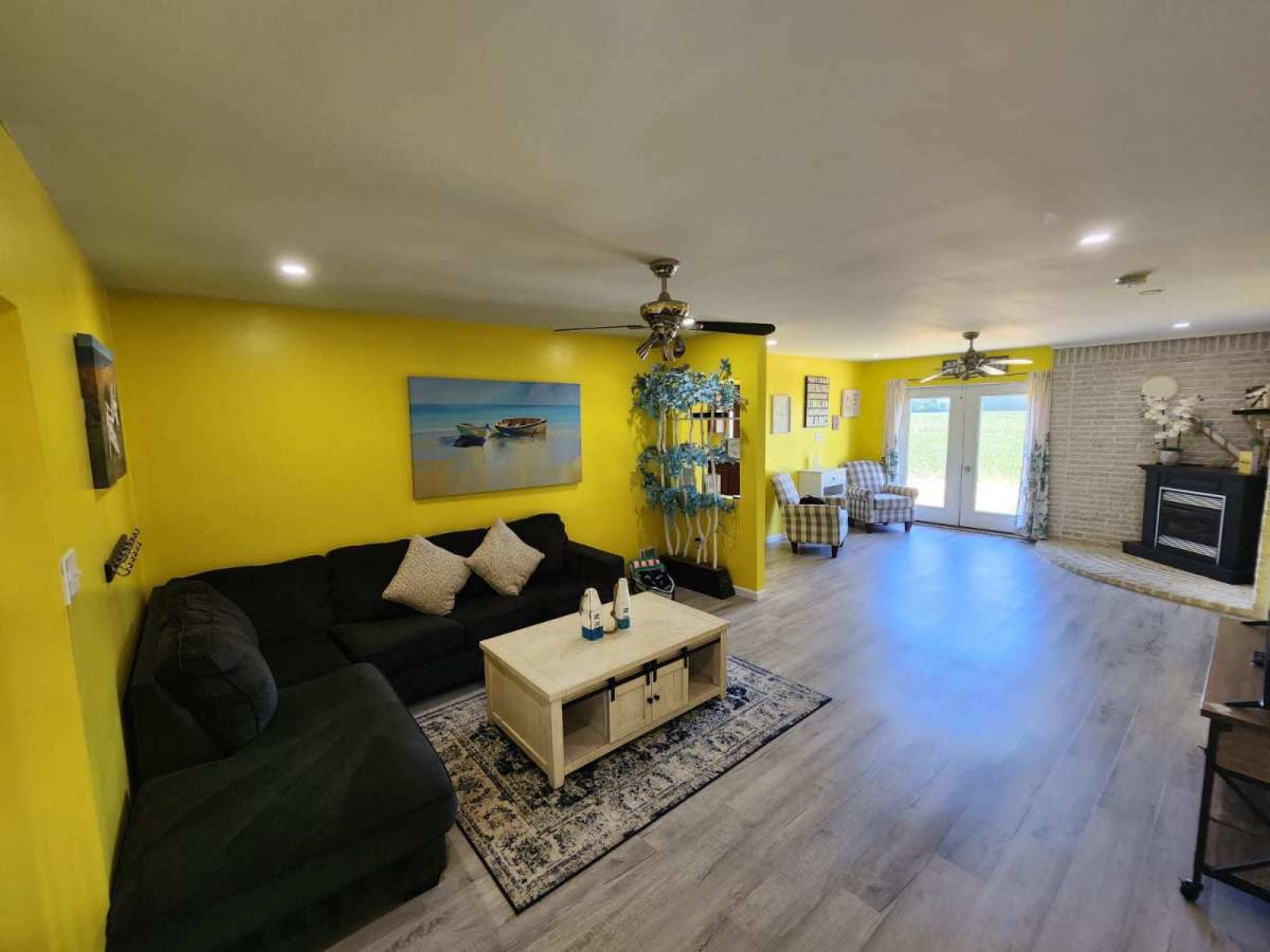 The living room features a dark sectional sofa, a coffee table, and a pair of armchairs, all set against bright yellow walls and a fireplace in the background.