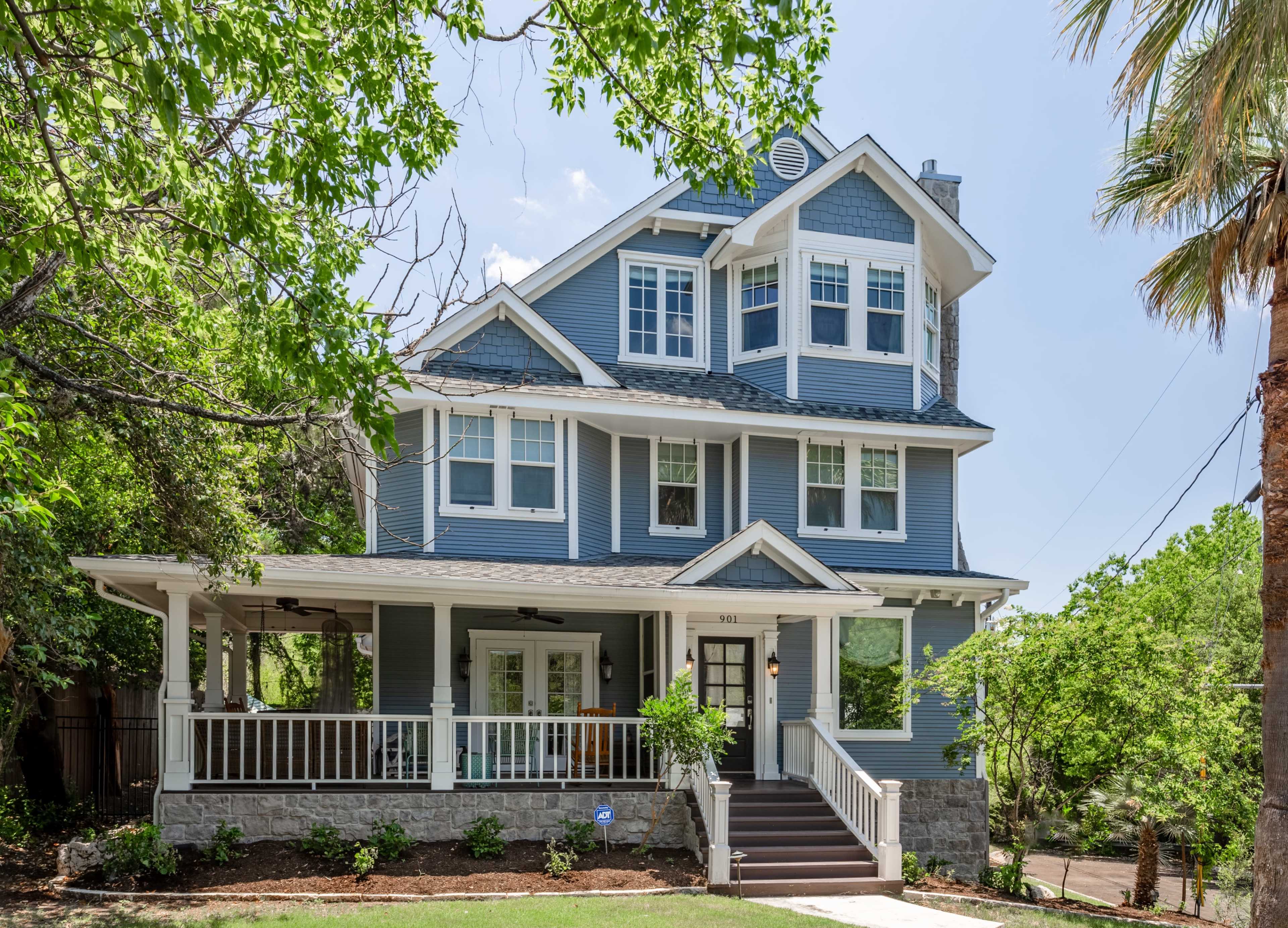 A large, two-story blue house with a wraparound porch and multiple windows surrounded by lush greenery.