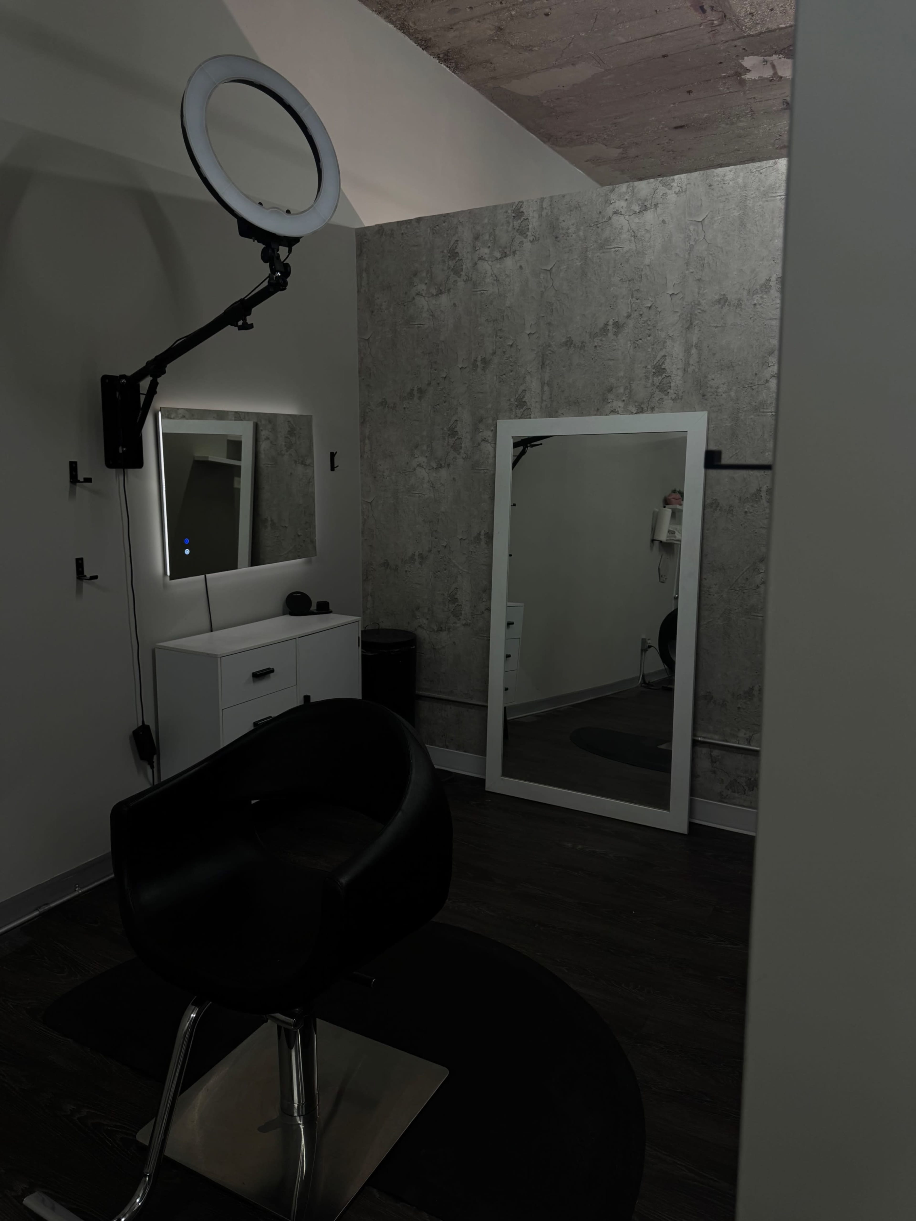 The image shows a minimalist salon room with a black chair, a large mirror, and a round light fixture mounted on the wall.