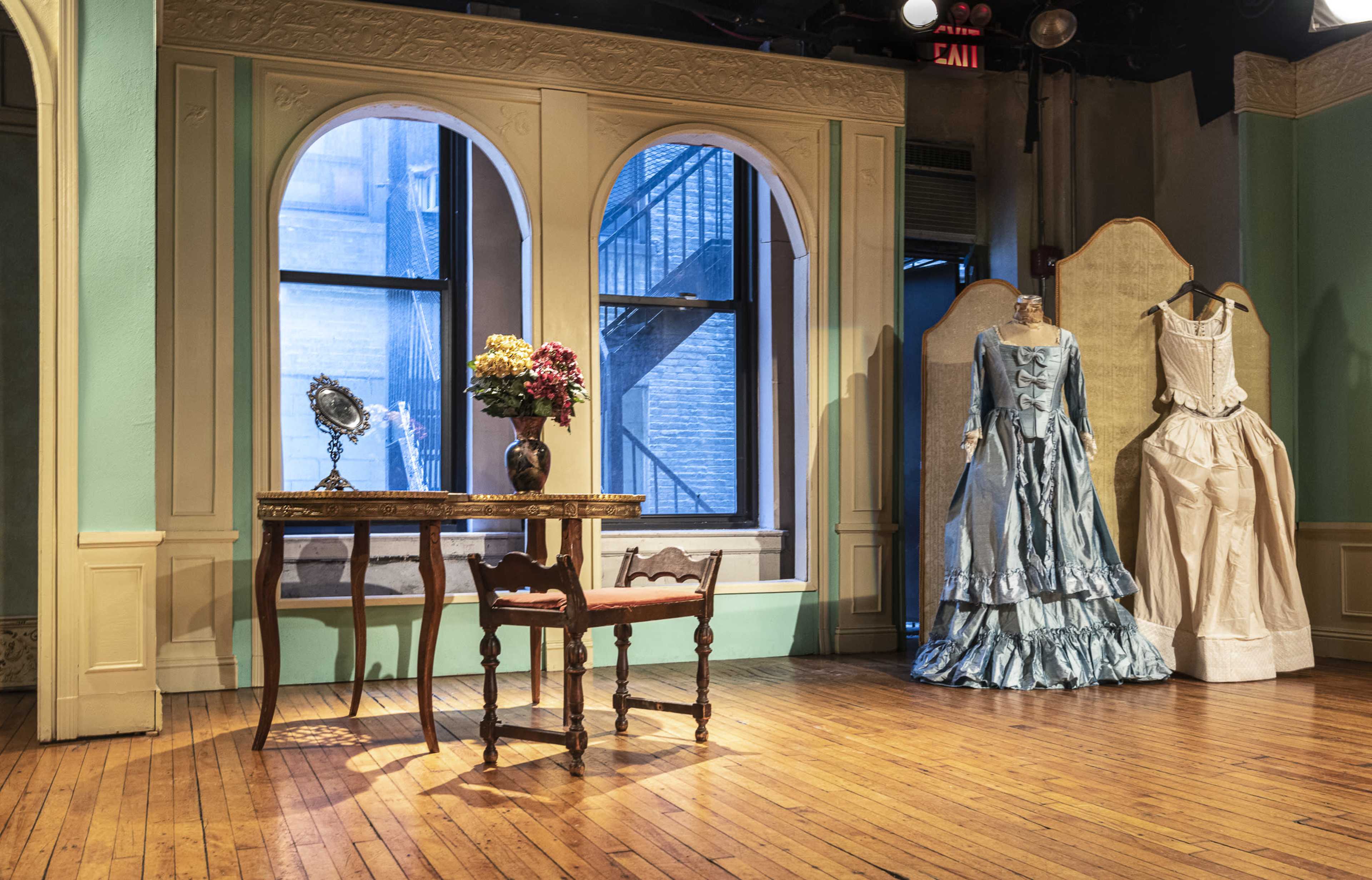 A room with wooden flooring features a table and chair, a vase of flowers, and two mannequins dressed in historical gowns, with large arched windows in the background.