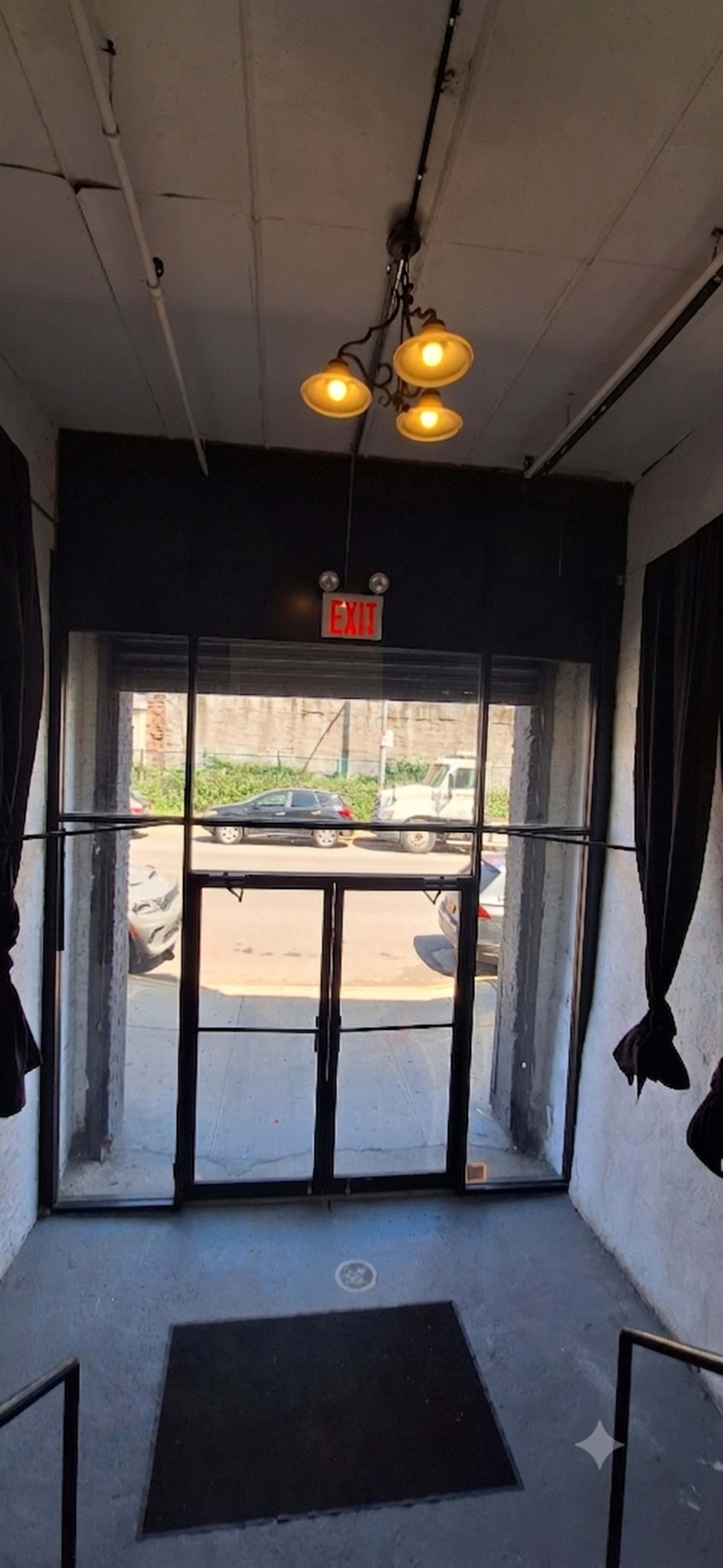 The image shows an interior entrance with a set of double glass doors leading to an outdoor area, flanked by black curtains and illuminated by a ceiling light fixture.