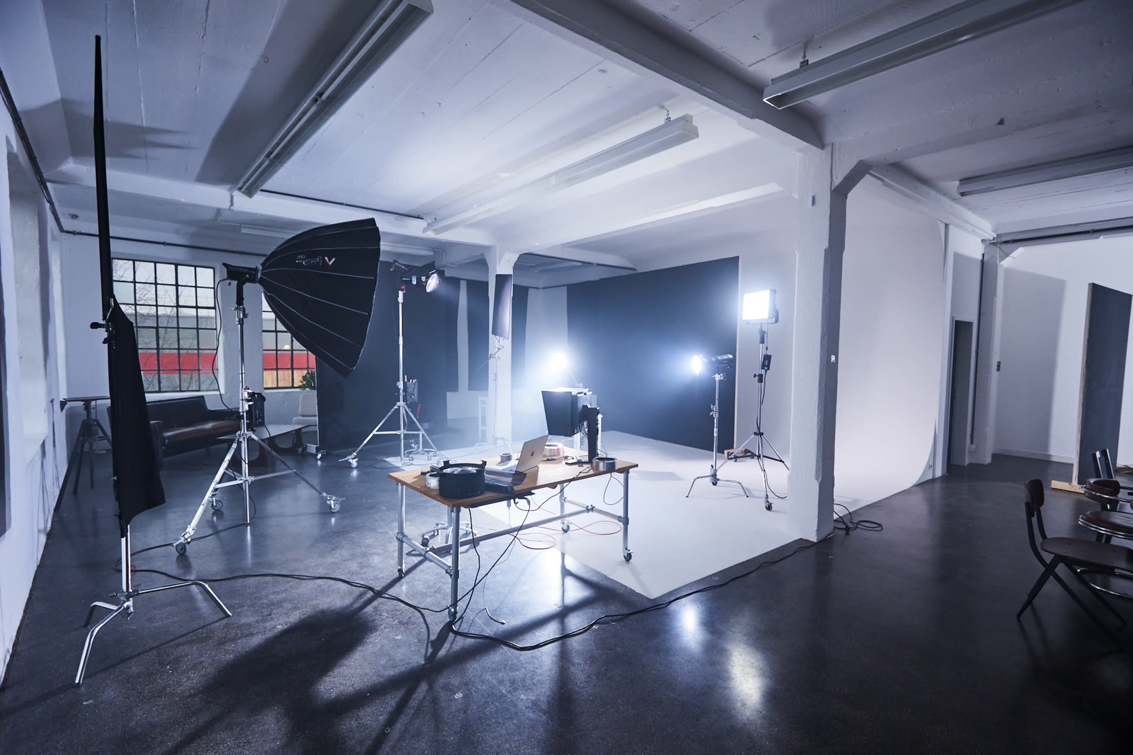 The image shows a well-lit photography studio featuring a central wooden table with equipment, a backdrop, and various lighting setups.