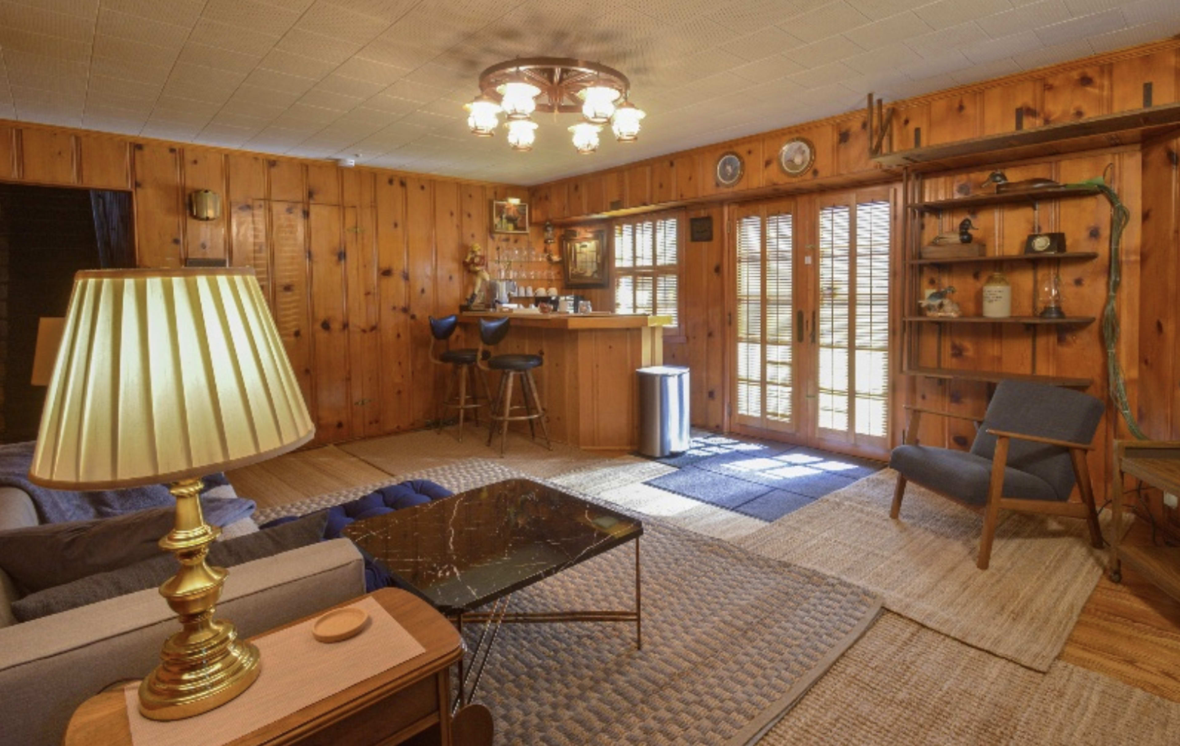 The room features wooden paneled walls, a bar area with stools, a coffee table, and a lamp next to a sofa, with sliding glass doors leading to an outdoor space.