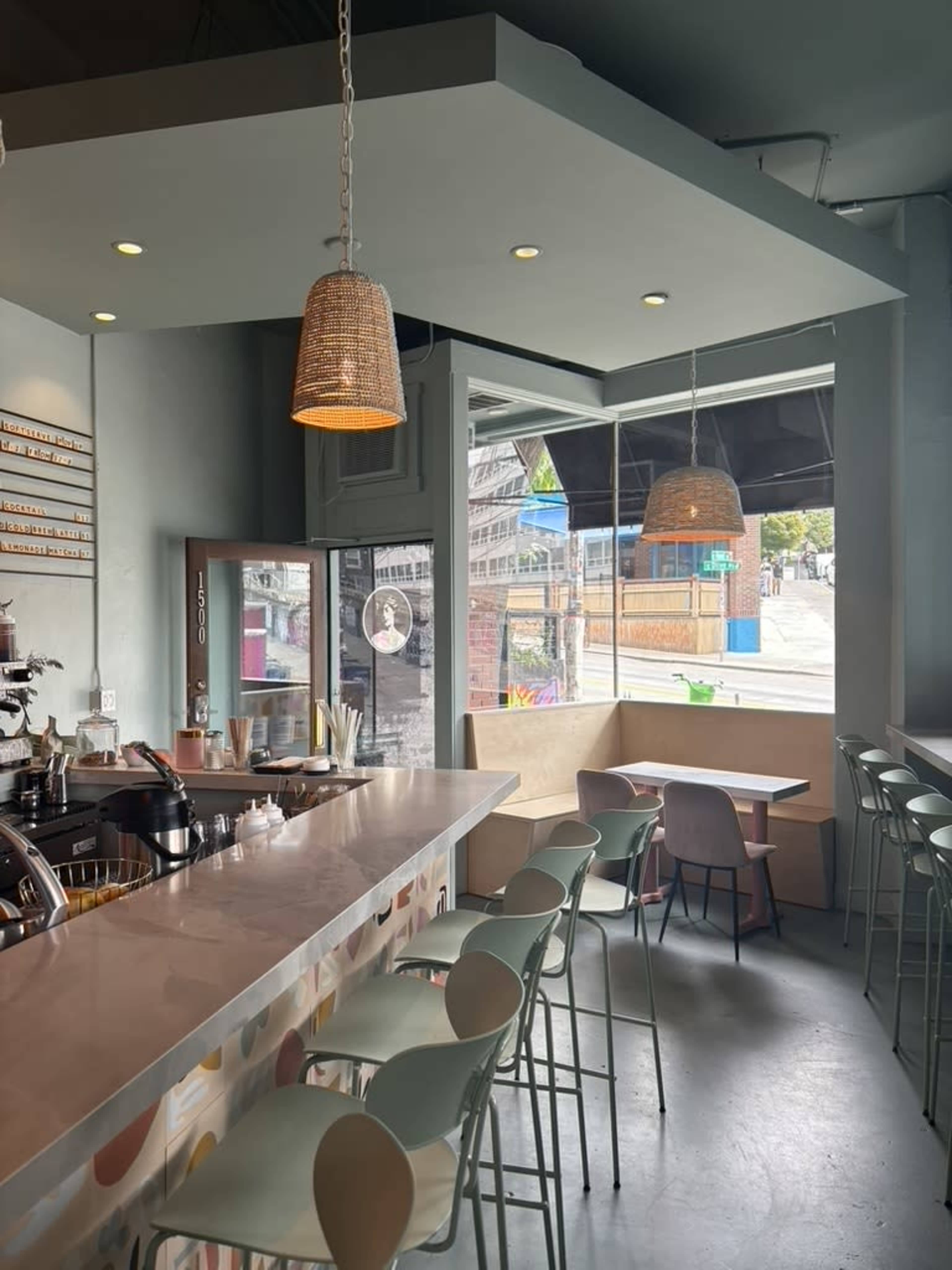The image shows a cozy café interior with a counter, bar stools, and large windows that provide a view of an outdoor area.
