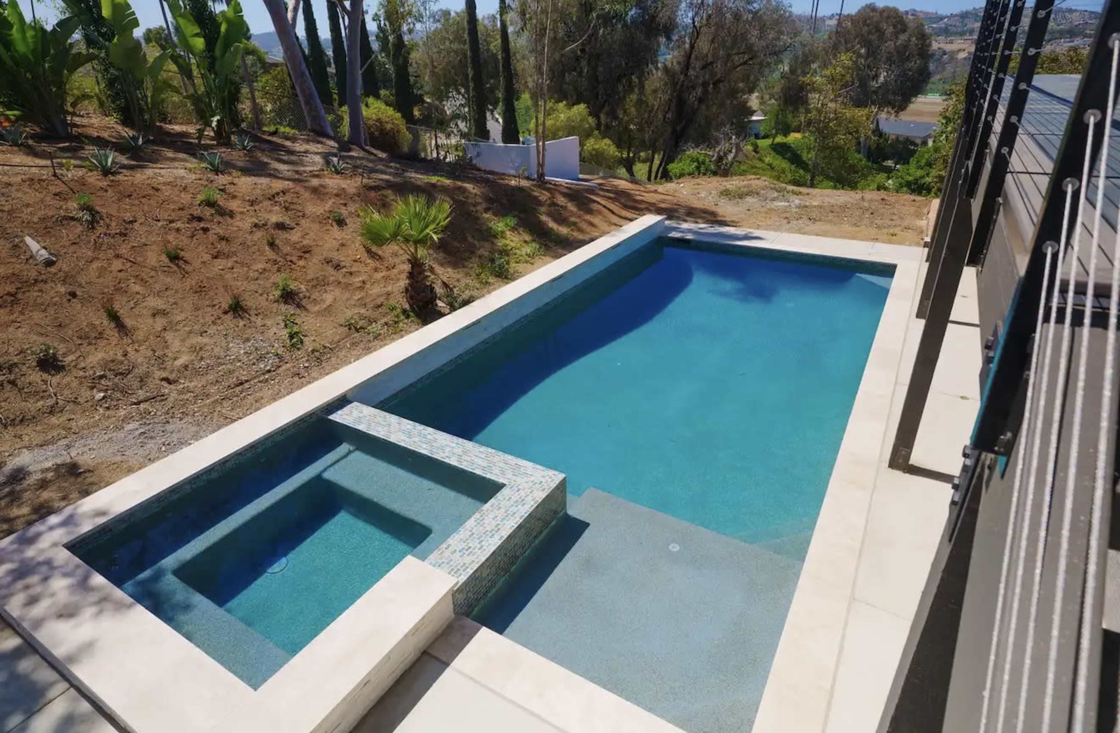 The image shows a rectangular swimming pool with a connected spa, surrounded by landscaped greenery on a slope.