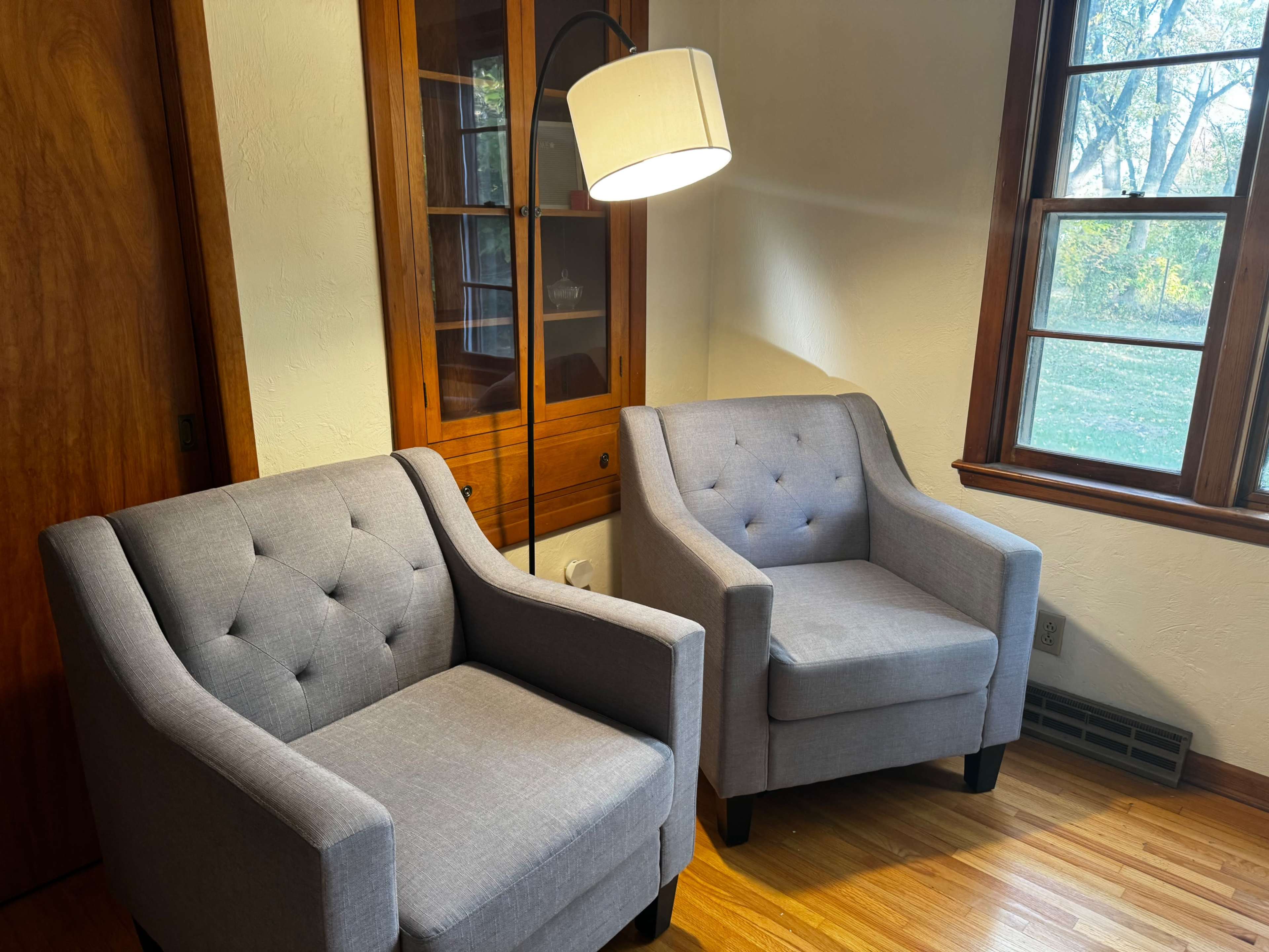Two gray upholstered armchairs facing each other in a well-lit corner of a room, with a floor lamp and a wooden cabinet in the background.
