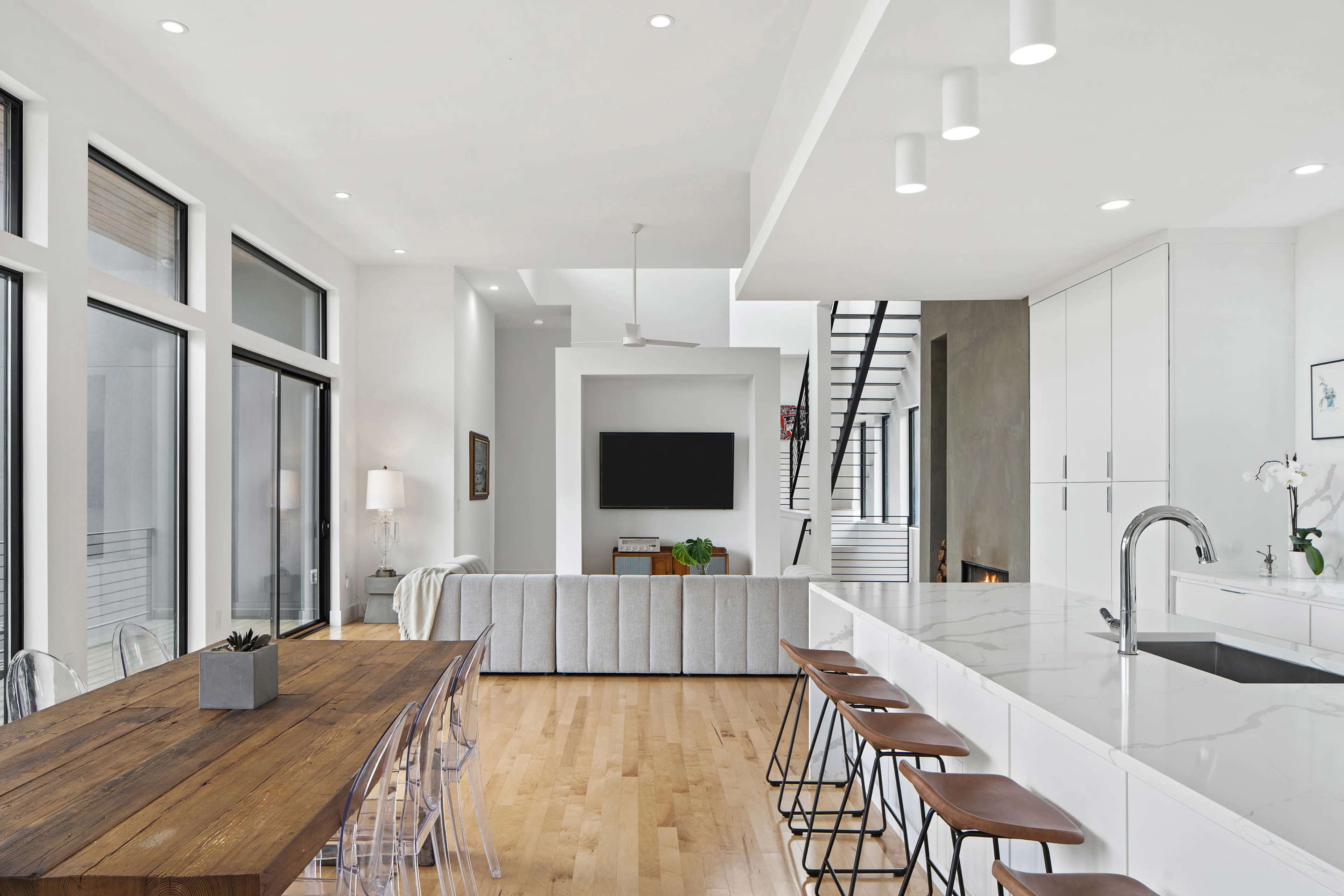 A modern open-concept kitchen and dining area features a large wooden table with transparent chairs, sleek cabinetry, and a marble countertop.