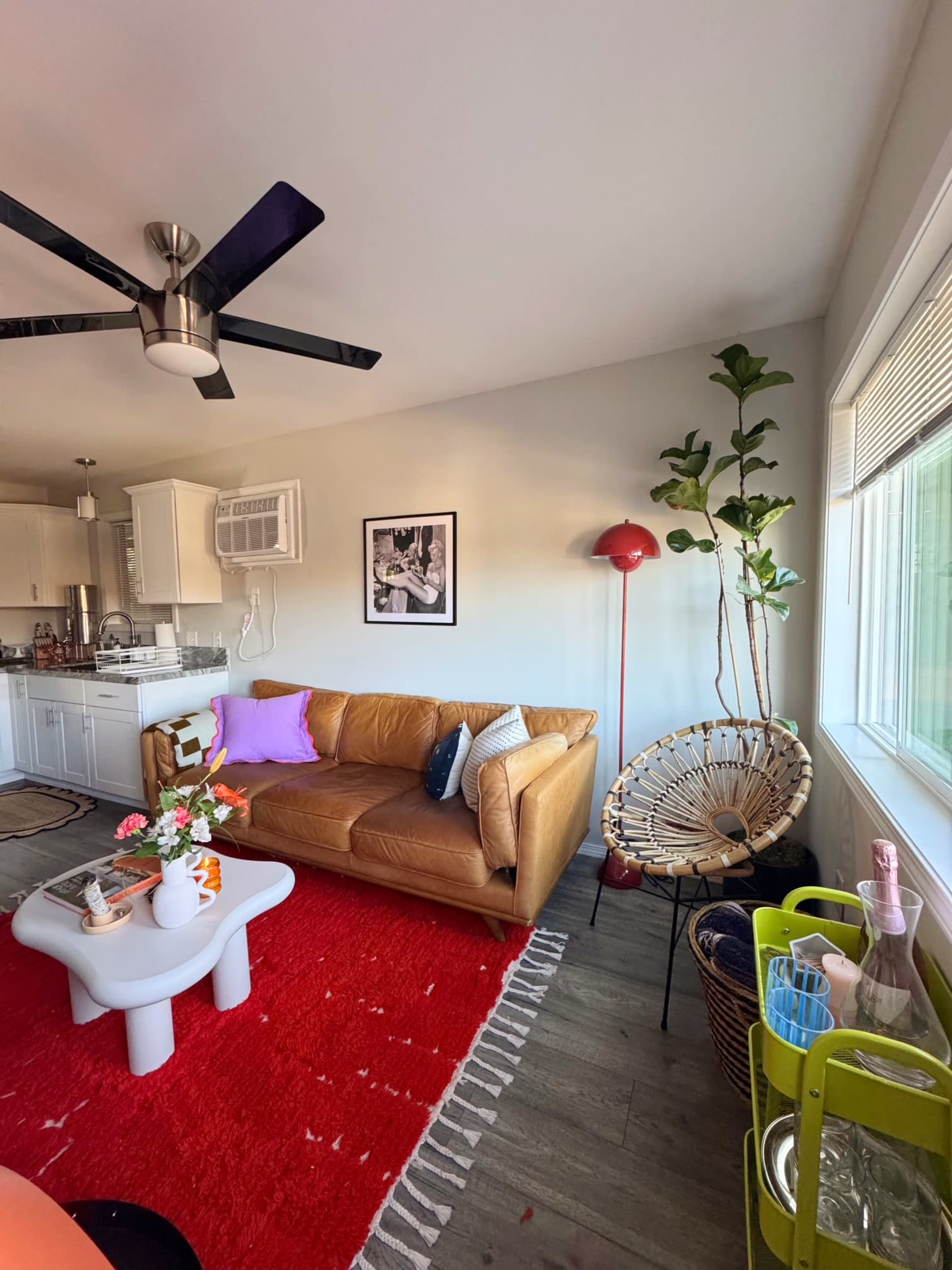 The image shows a modern living room featuring a brown leather sofa, a white coffee table, and a vibrant red area rug, accompanied by a tall plant and various decorative items.