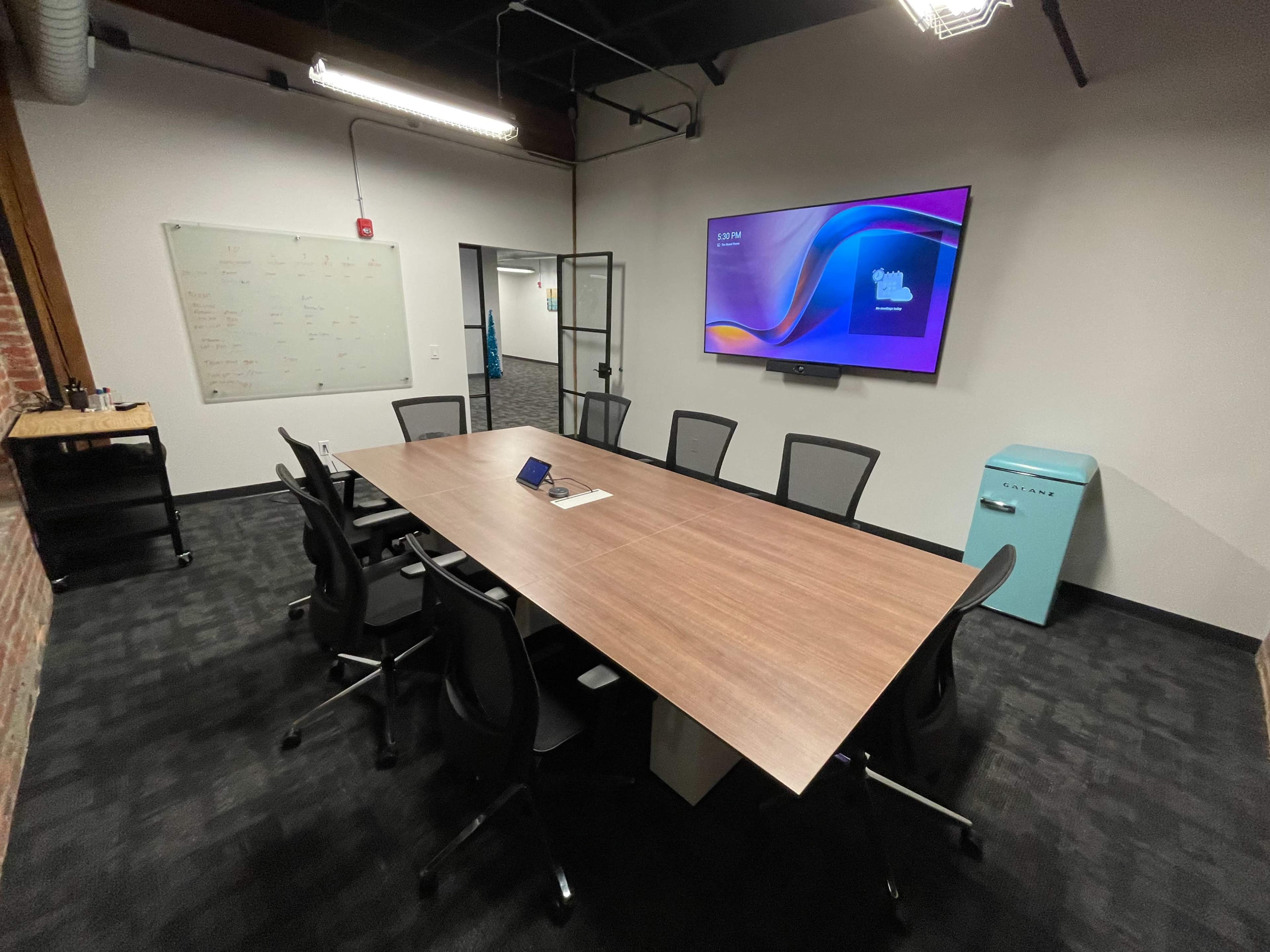 A modern conference room features a large wooden table surrounded by black chairs, a wall-mounted screen displaying a presentation, a glass door, a whiteboard, and a retro-style blue refrigerator.