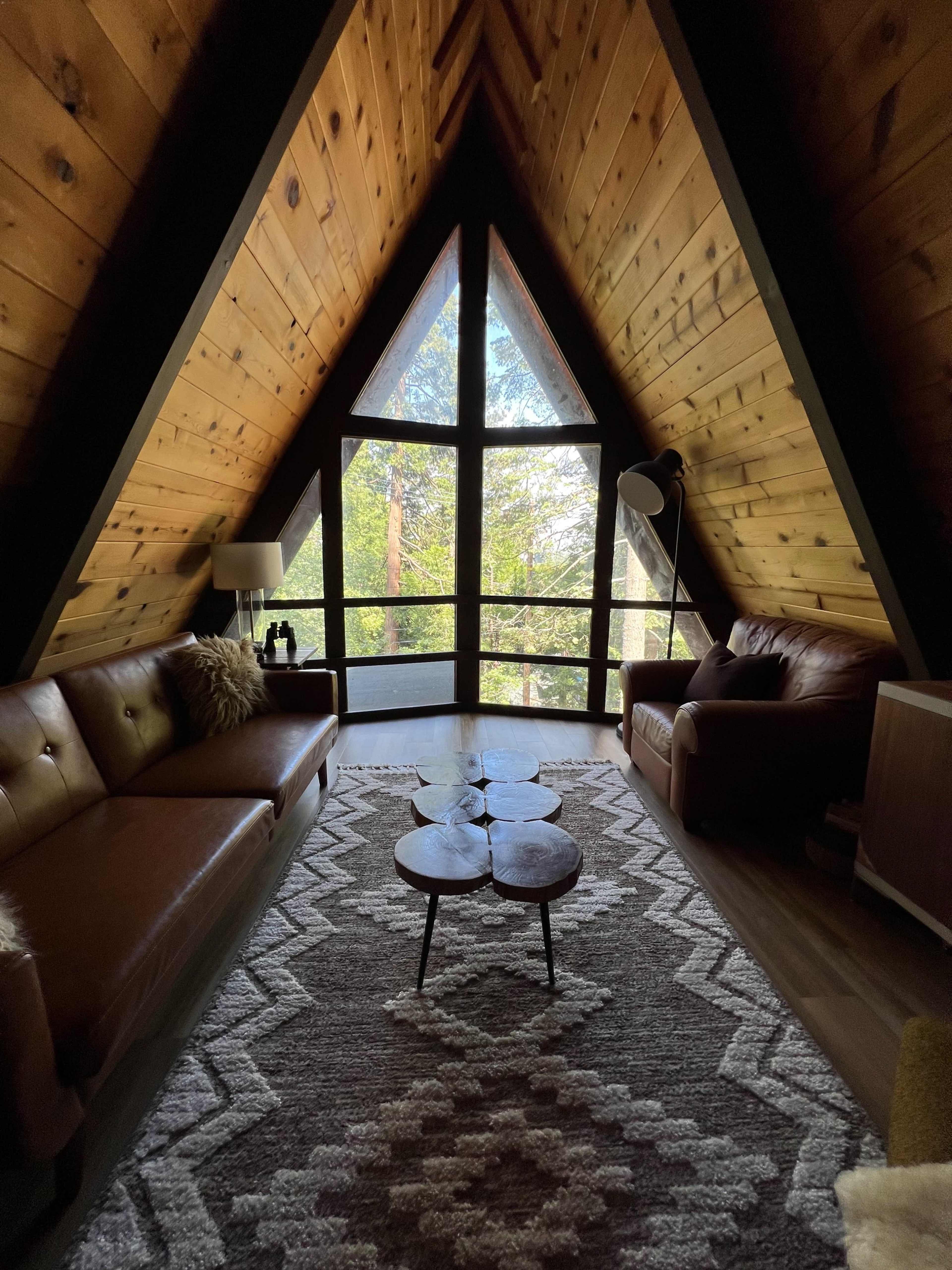 The interior of an A-frame cabin features wooden walls, a large triangular window, two leather sofas, a patterned rug, and a coffee table made of three wooden slabs.